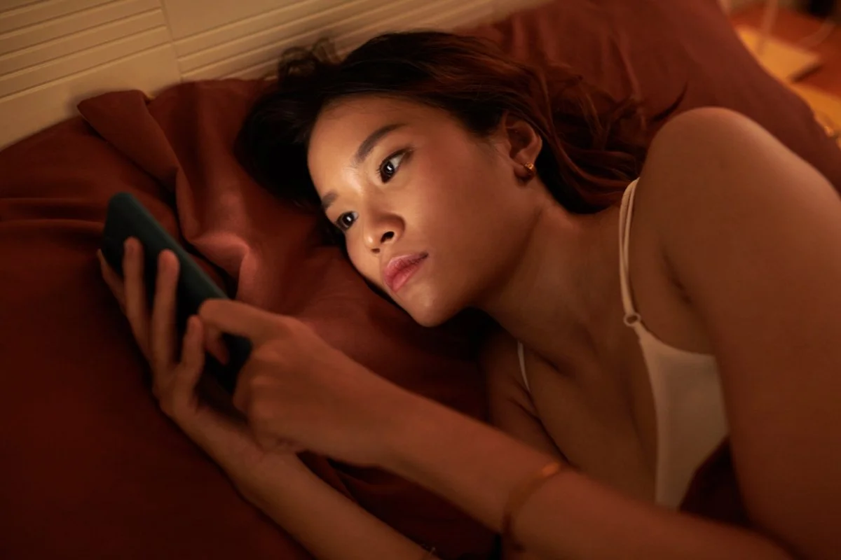 Woman lying in bed looking at her phone at night illustrating doomscrolling anxiety and difficulty mentally disconnecting before sleep