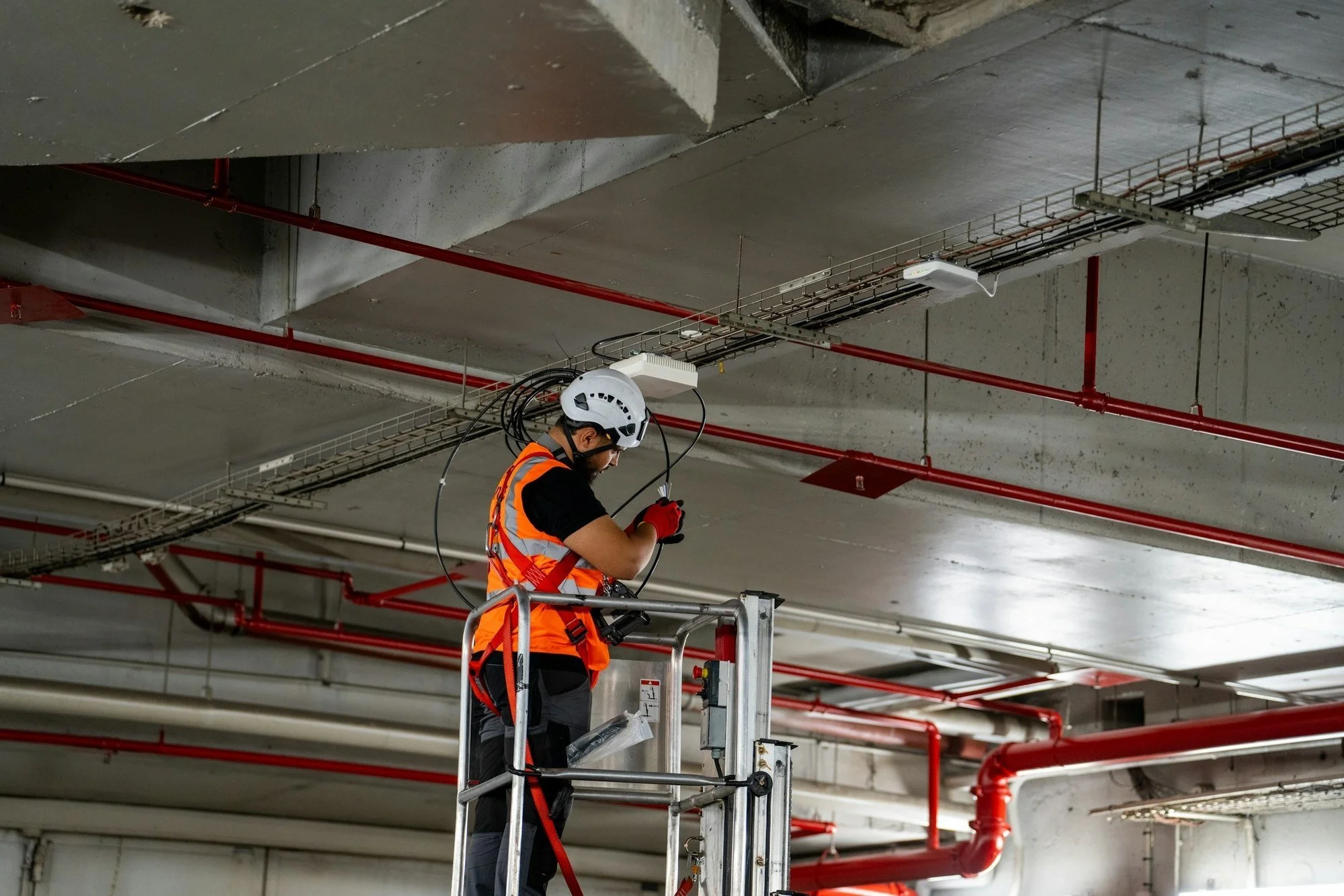 Electrician in a hard hat and harness on a lift installing cable tray beneath fire sprinkler piping on a mission-critical build