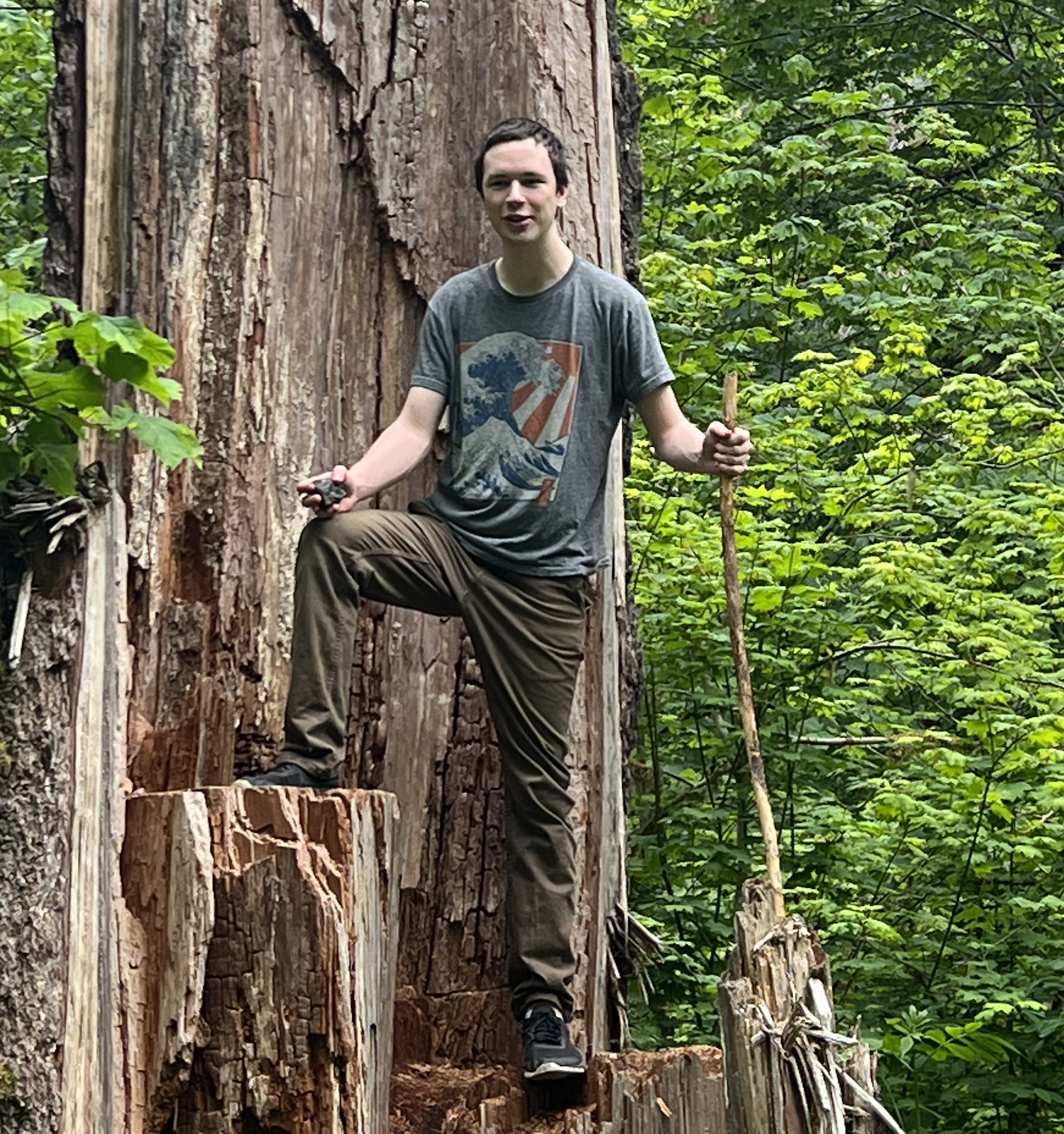 St. John Stephens holding a large walking stick, standing triumphantly on a tree stump.