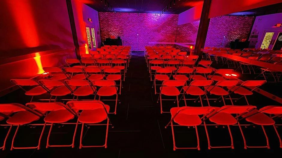 Rows of seating facing a microphone and performance area at an A.M.P. Open Mic gathering.