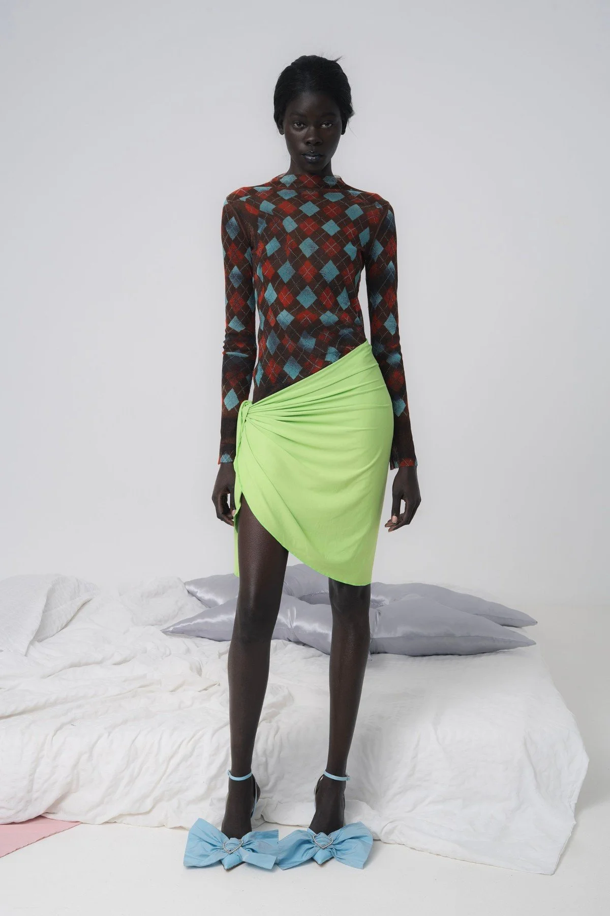 Model in an argyle mesh top and lime sarong skirt before a bed