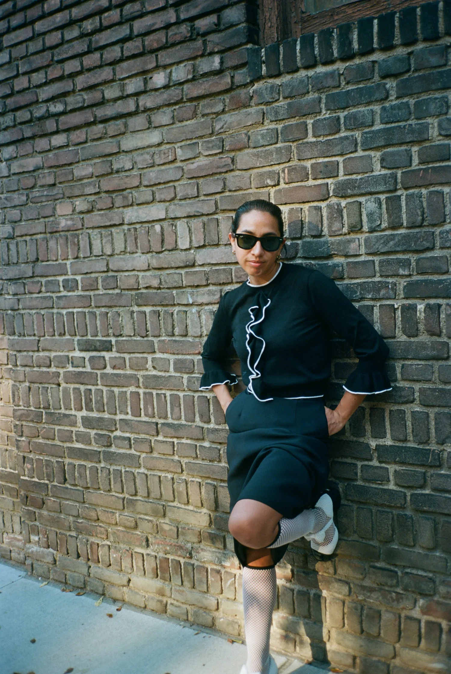 Malén Denis in sunglasses and a black ruffled top, leaning on a brick wall with one knee raised