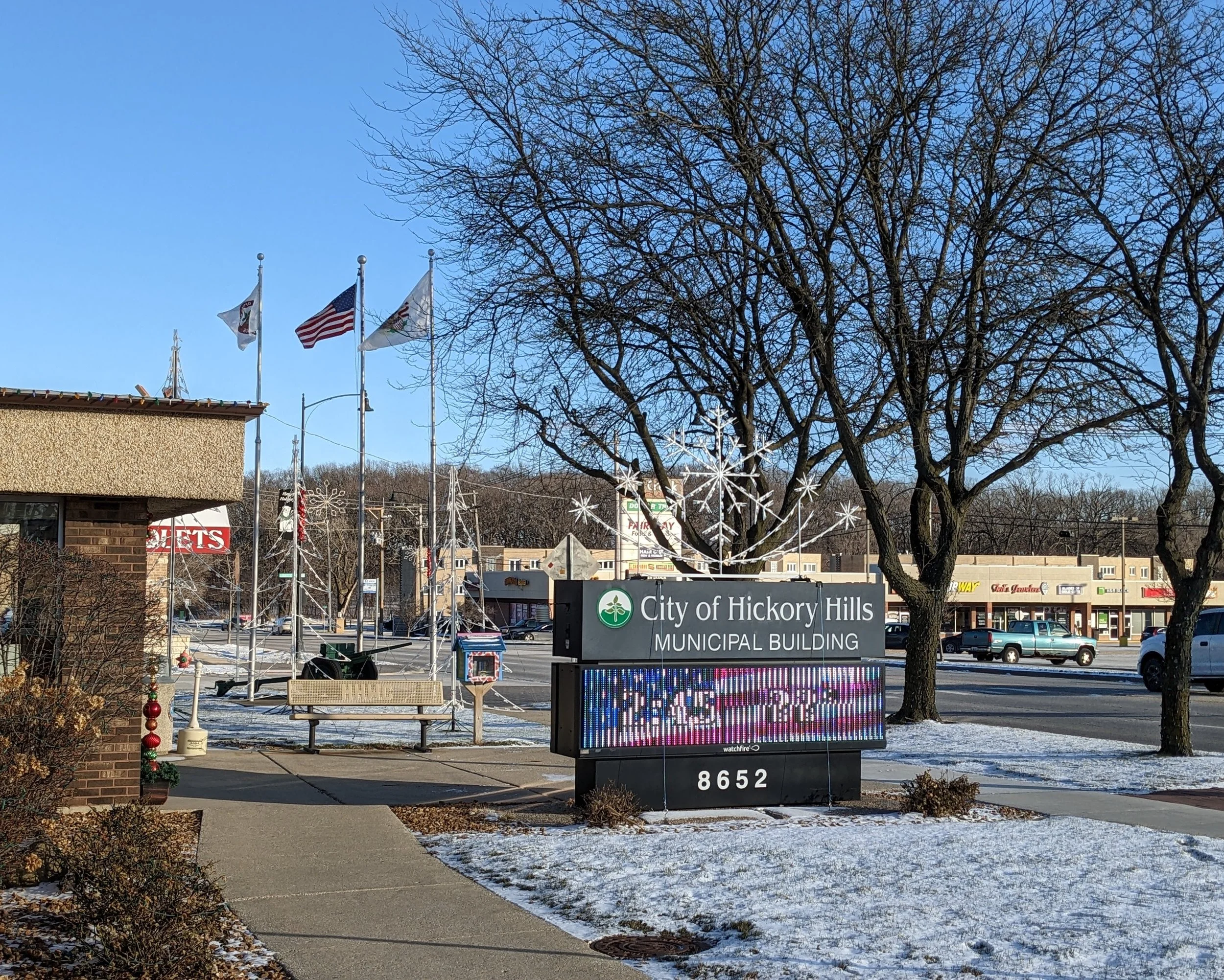 Flags and the City of Hickory Hills Municipal Building sign beside a snowy sidewalk
