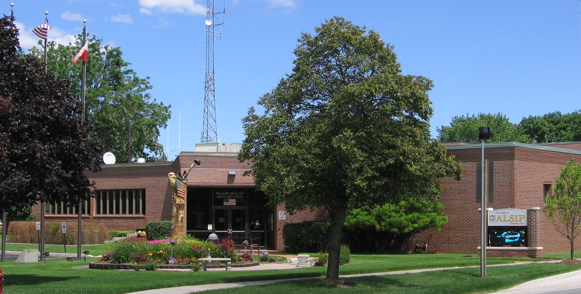 Brick Alsip Village Hall with flower beds, flagpoles, and a village sign in front