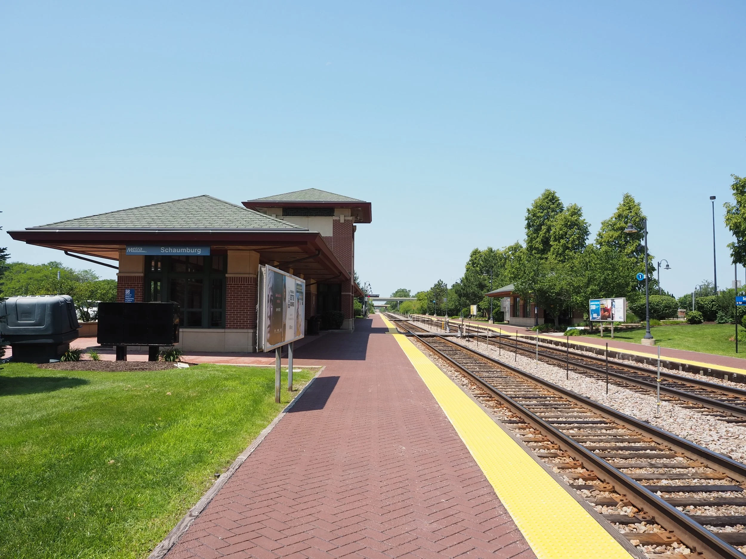 The brick Metra Schaumburg station building and its platform on a clear day, with the tracks running along the right side