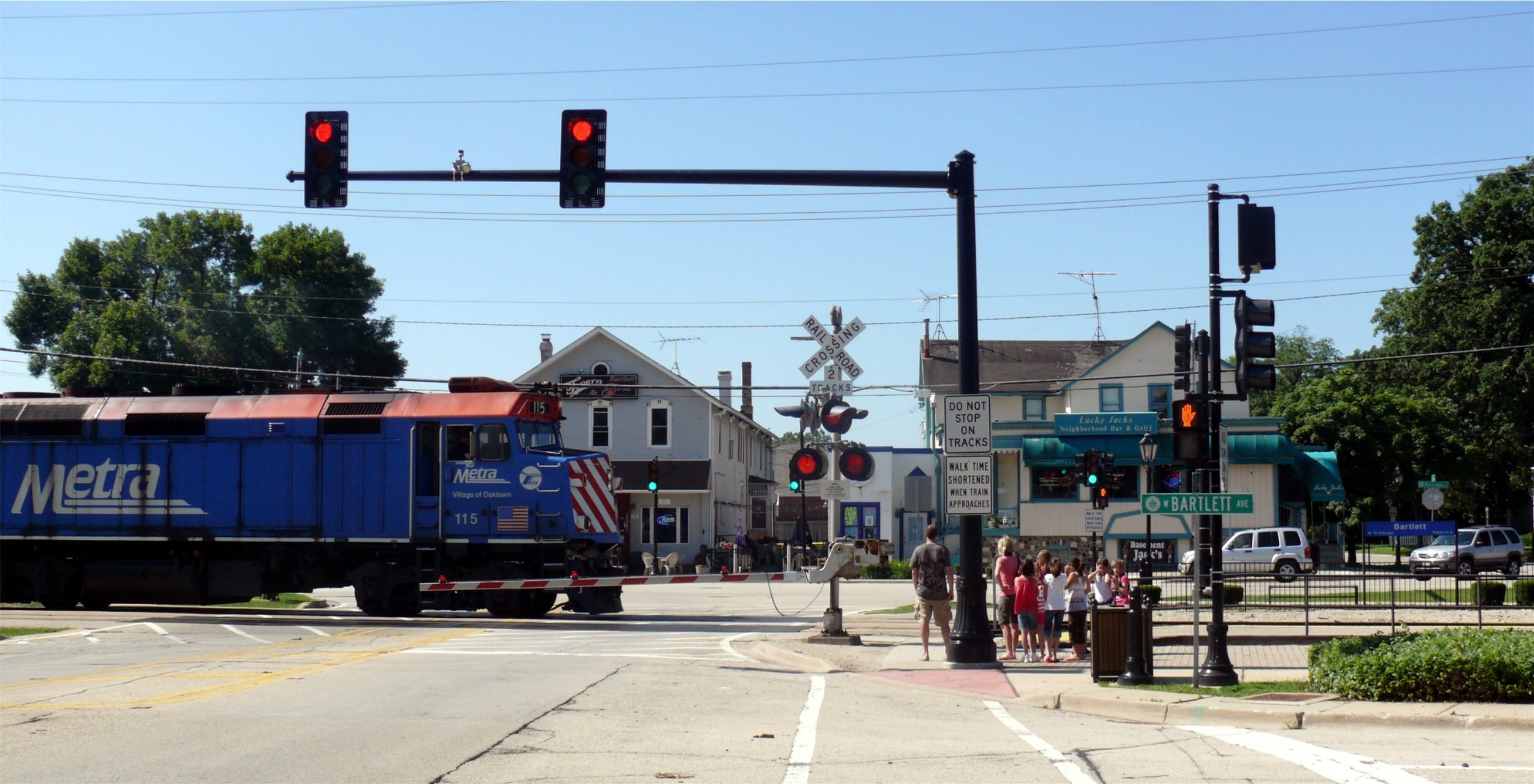 A Metra train stopped at the Bartlett Avenue crossing in downtown Bartlett, with older frame shop buildings along the street