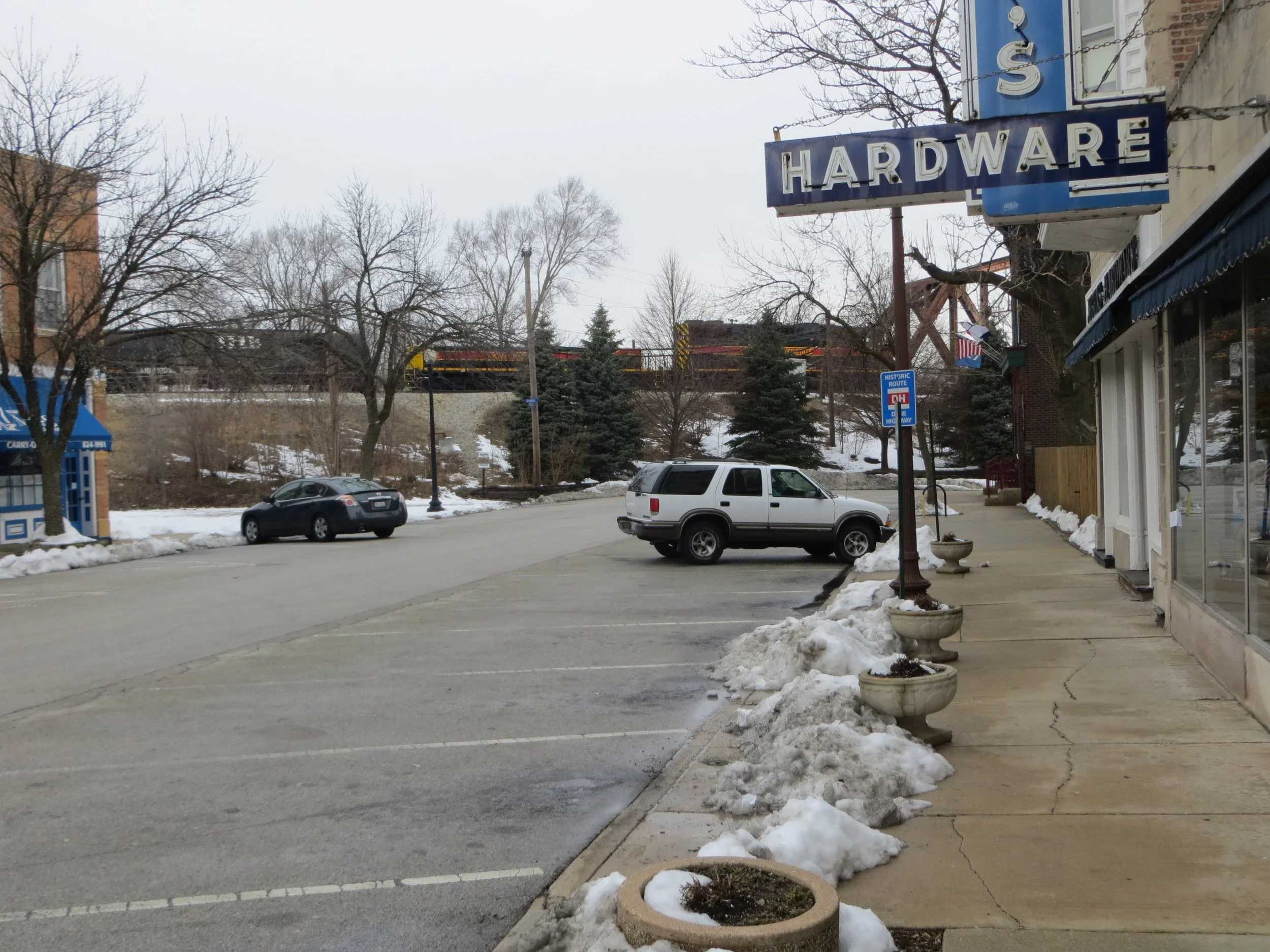 Snowy parking lane along Olde Western Avenue in Blue Island, with a hardware store sign and a freight train on an embankment behind