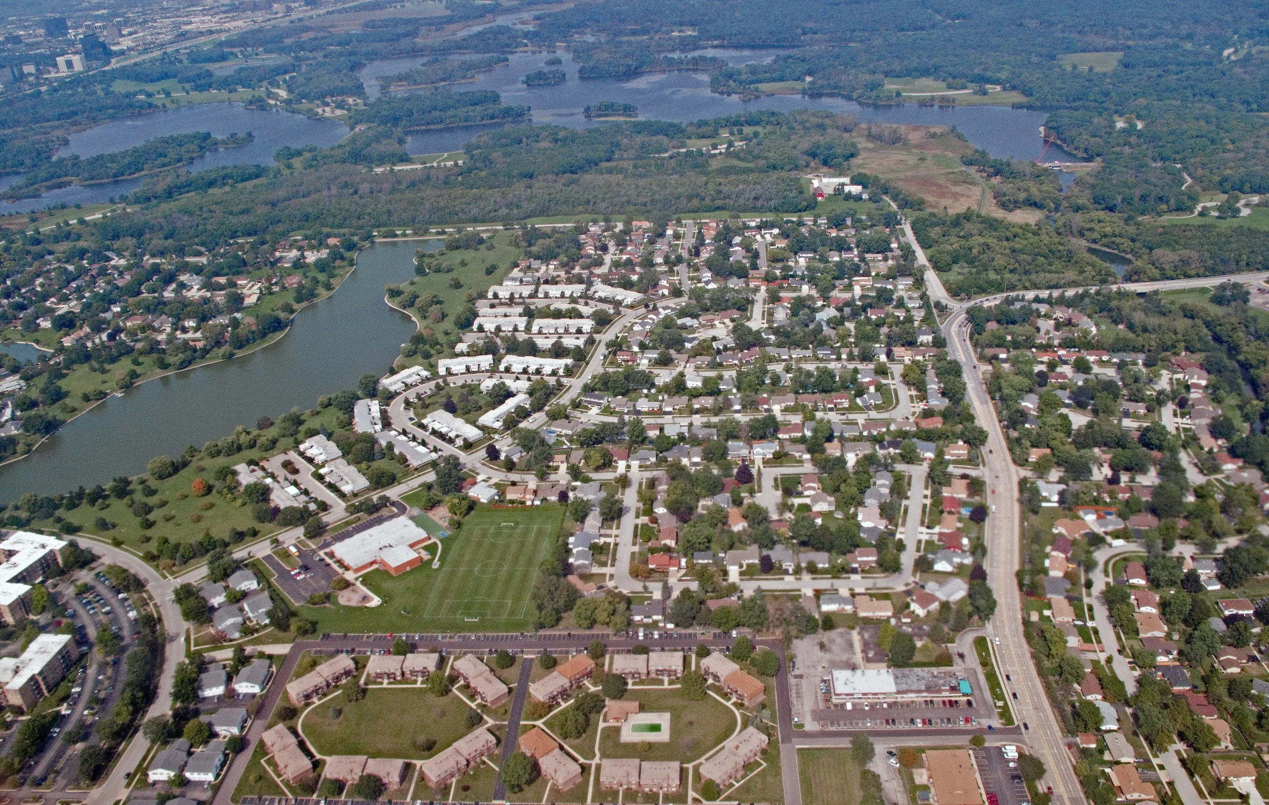 Aerial view of Elk Grove Village with a lake and the Busse Woods forest preserve above neighborhoods of houses, townhomes, and a soccer field