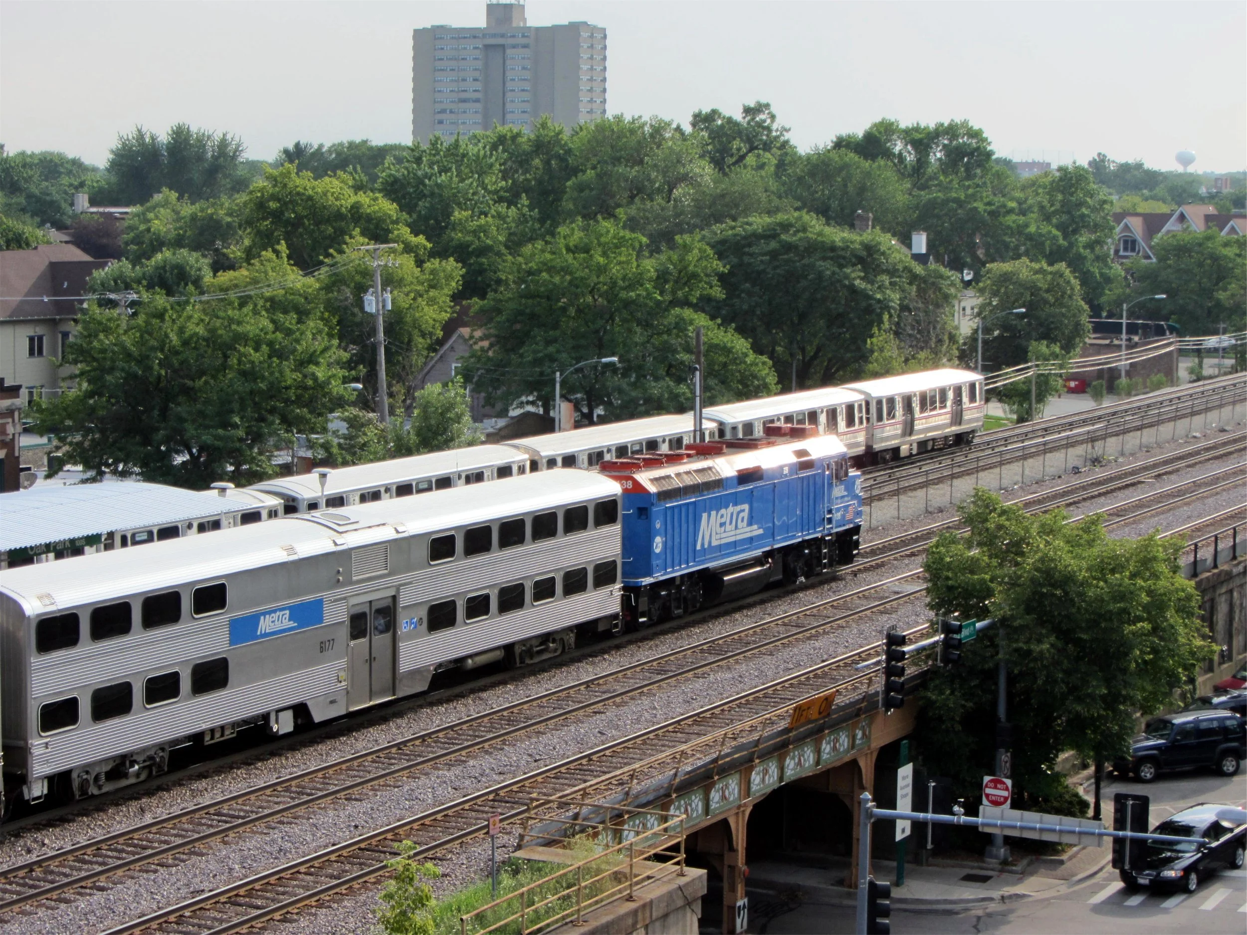 A blue Metra locomotive pulling bilevel cars past a CTA train in Oak Park, with tree-lined blocks of houses behind the tracks