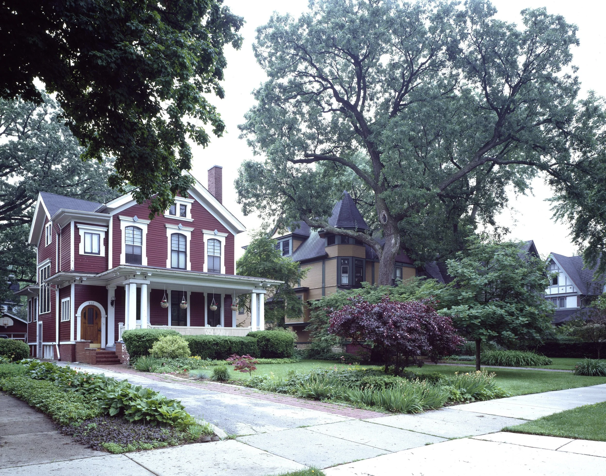 A red frame house with a deep front porch beside a house with a pointed tower, under tall trees on an Oak Park street