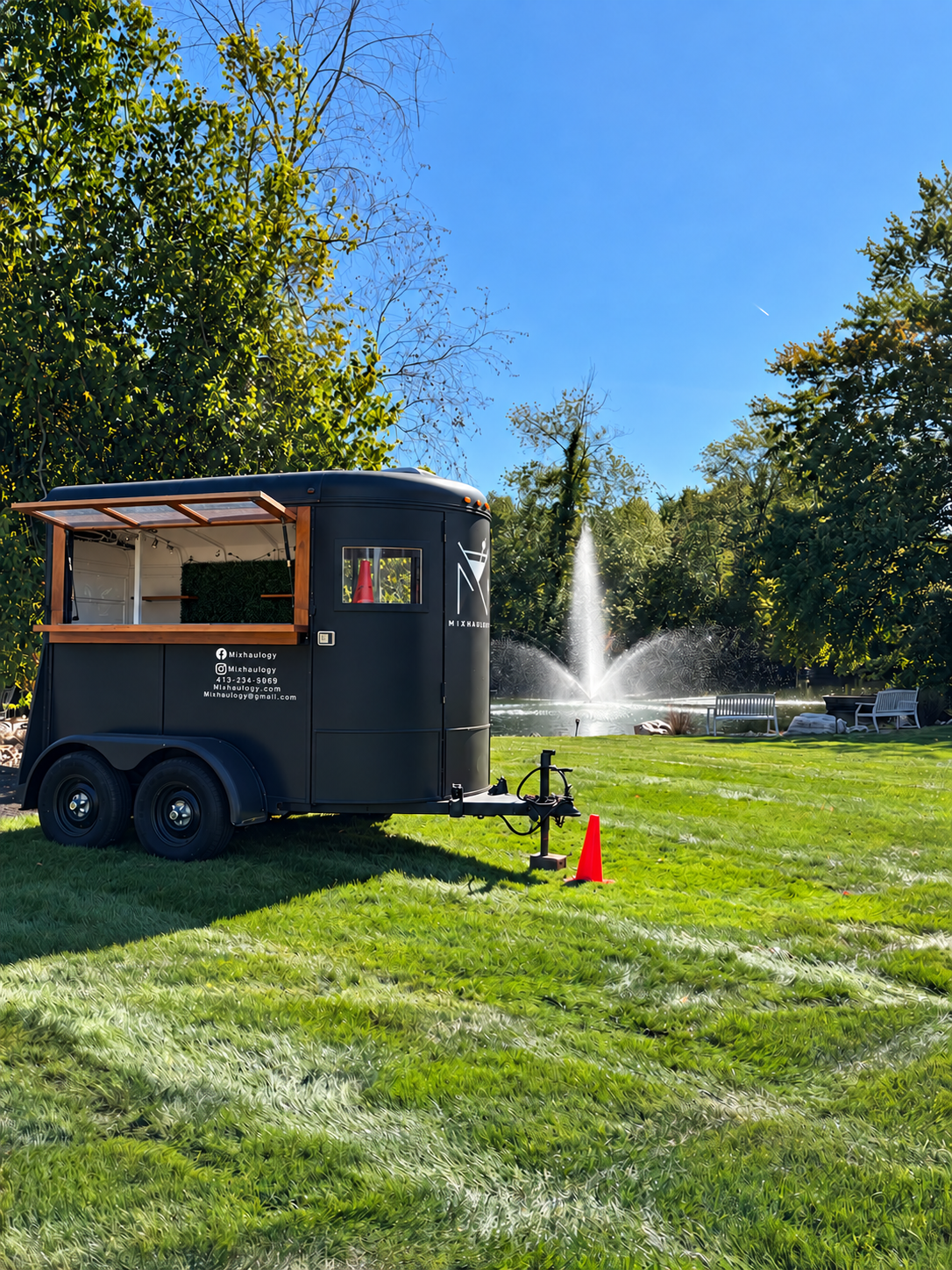 Mixhaulogy small mobile bar trailer beside a pond and fountain, digitally edited to show the serving window open