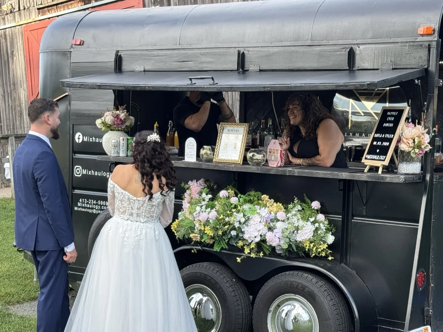 A bride and groom chatting with Mixhaulogy bartenders at a flower-decorated mobile bar