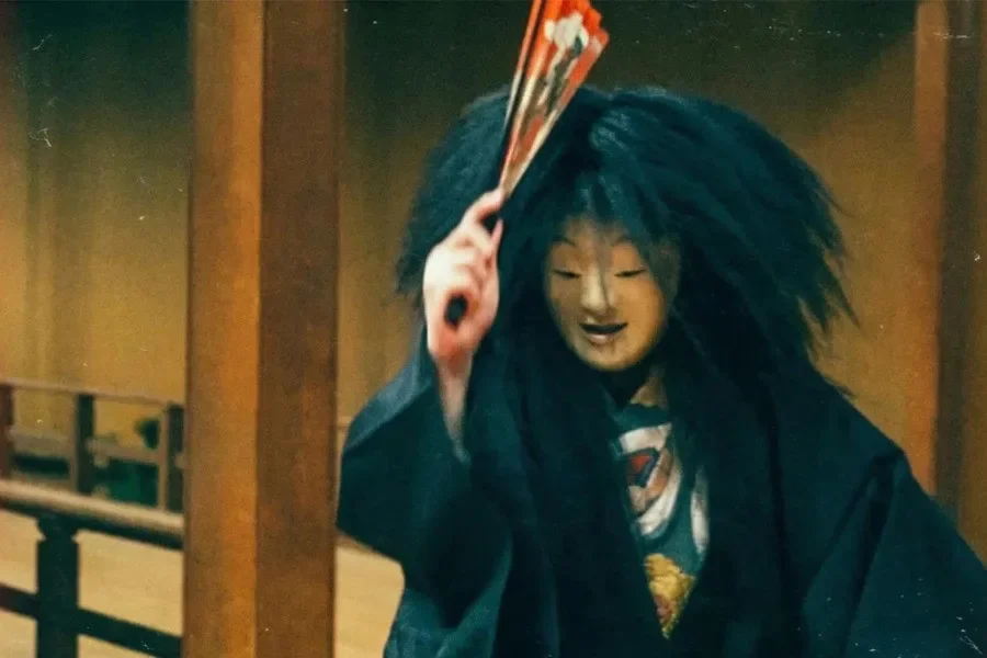 A Noh performer in a pale mask and long black wig raising a red fan on a wooden stage