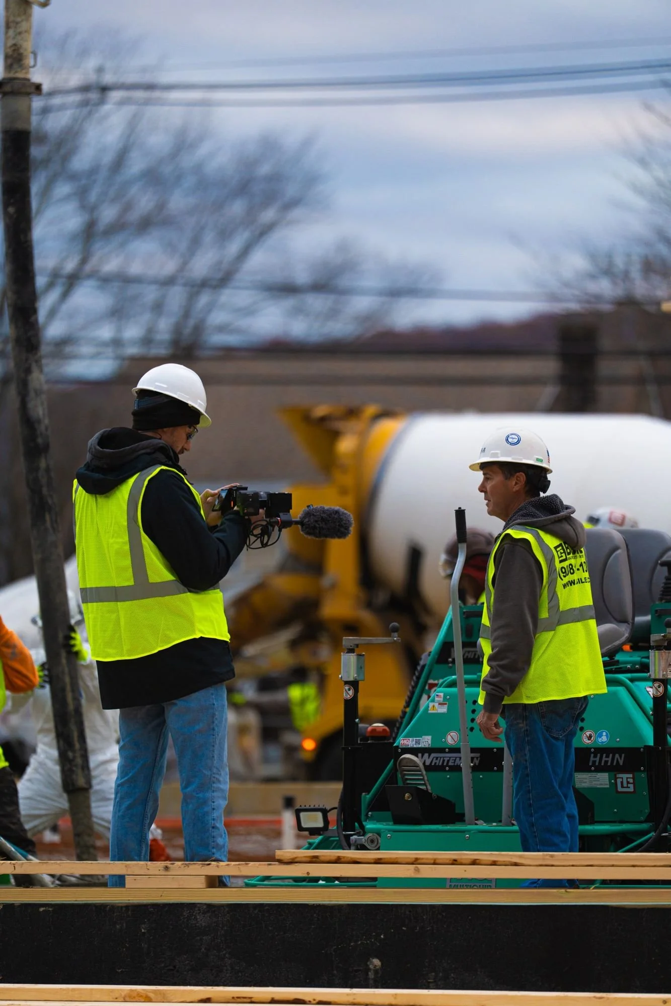 Camera operator in hi-vis filming a concrete pour with a pump truck behind