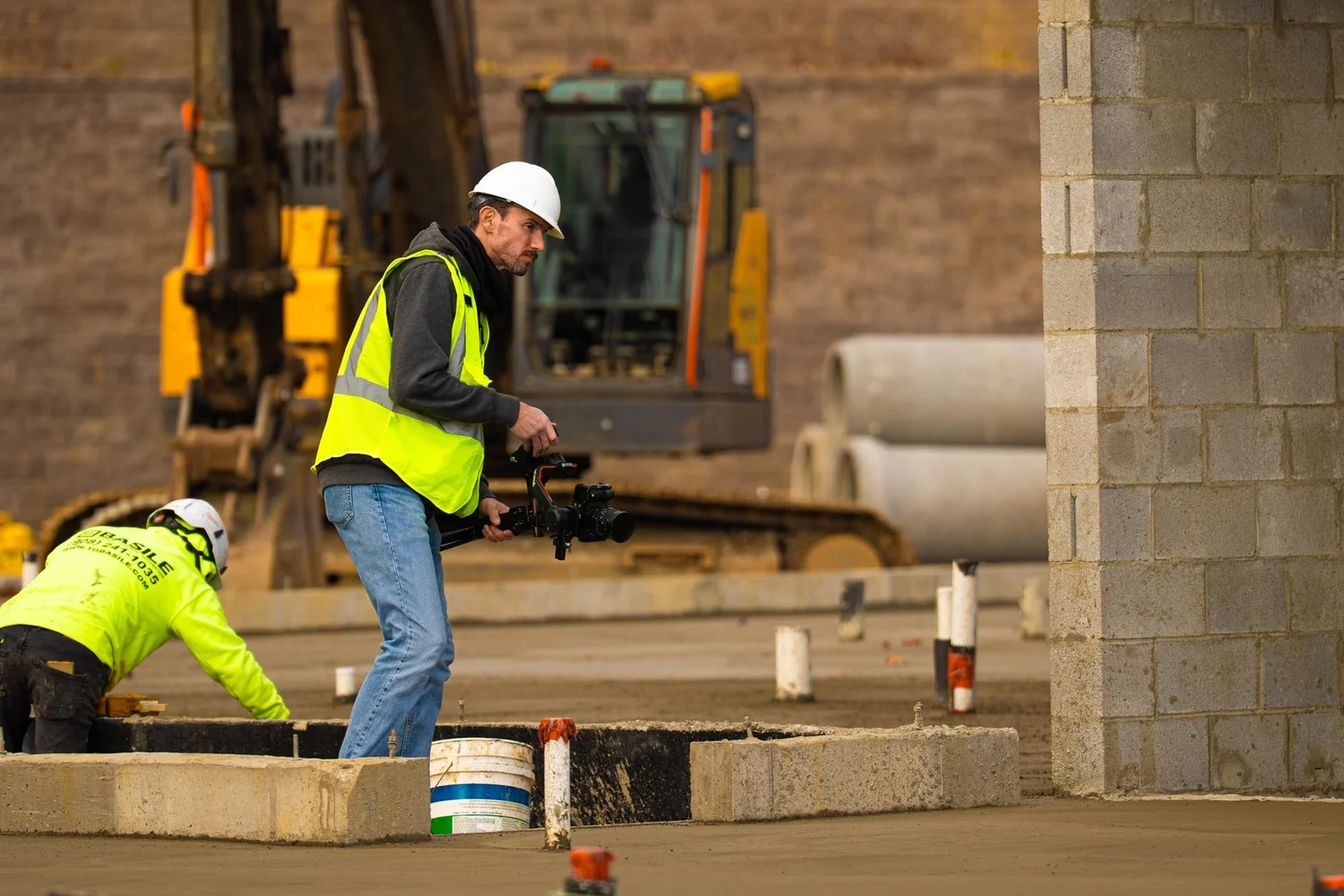 Videographer filming a mason at a block wall on a jobsite, excavator in the background