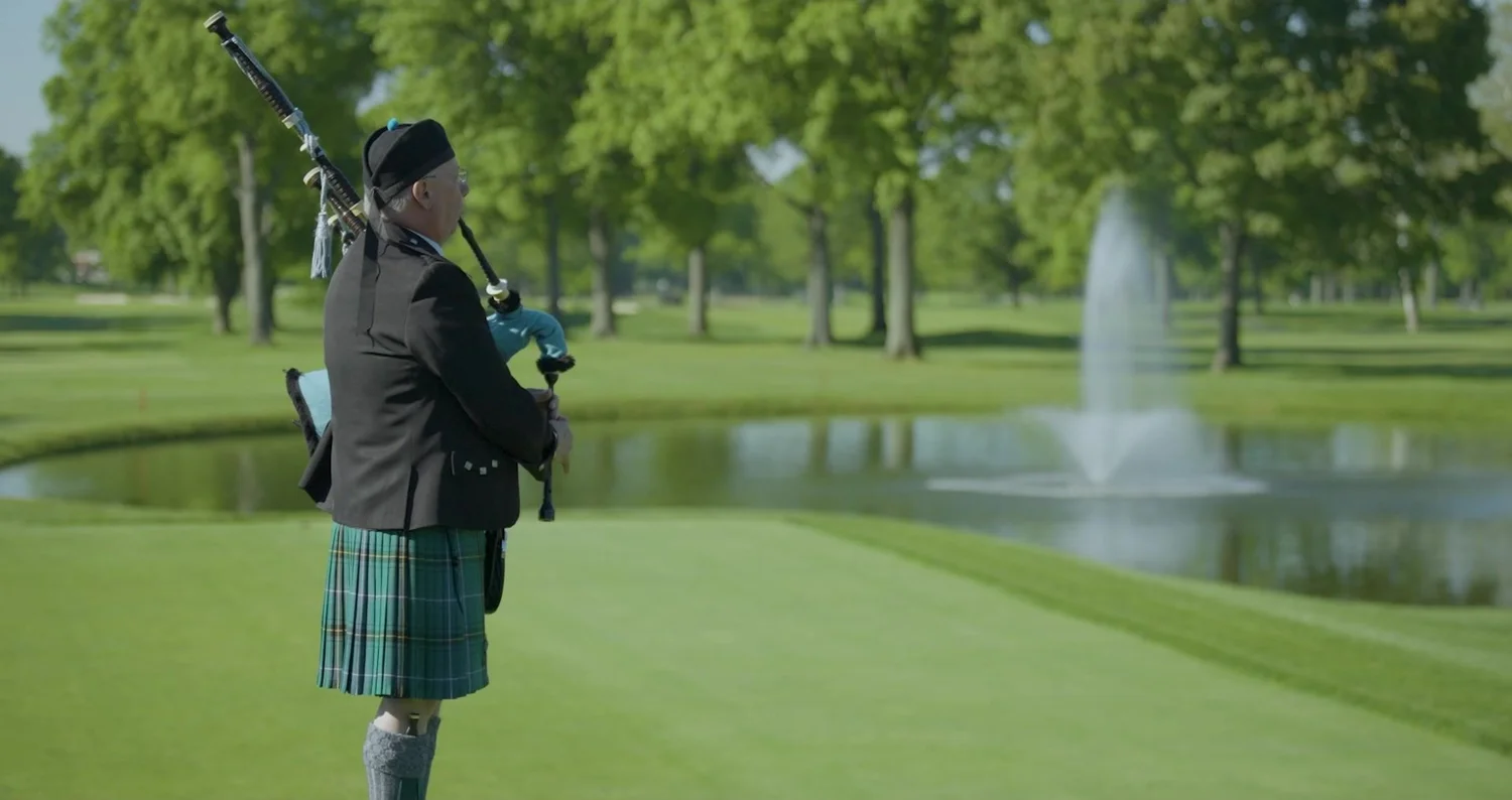 Bagpiper in a kilt beside a pond and fountain on a private golf course