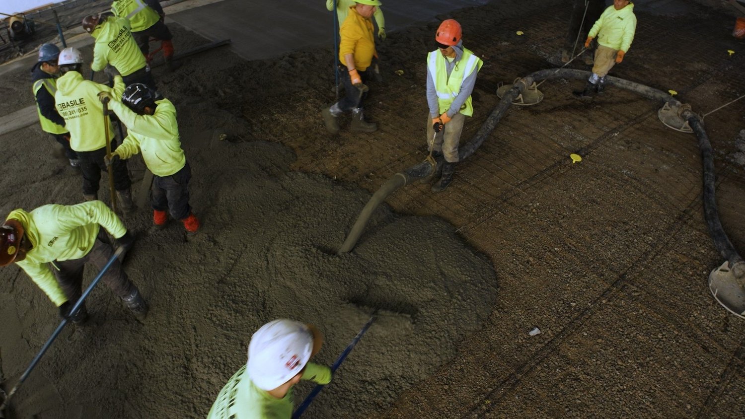 Overhead construction progress video frame of a crew in hi-vis placing and raking concrete