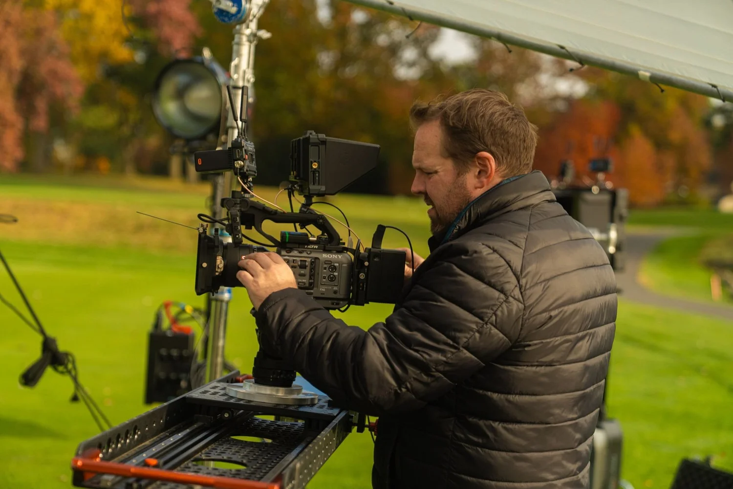 Camera operator framing a shot on a cinema rig at a private club, fall trees behind