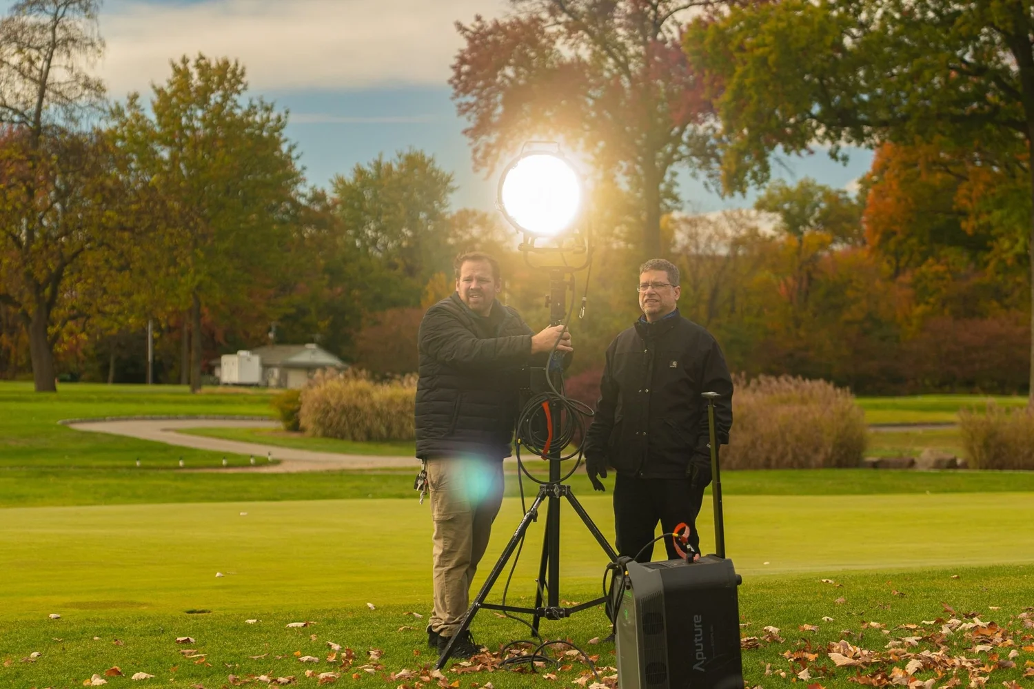 Film crew backlit by a low sun beside a light stand on a golf course