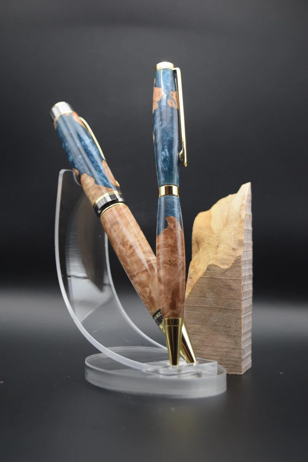 Two handmade pens of burl wood and blue resin on a clear stand, beside the raw blank they were turned from