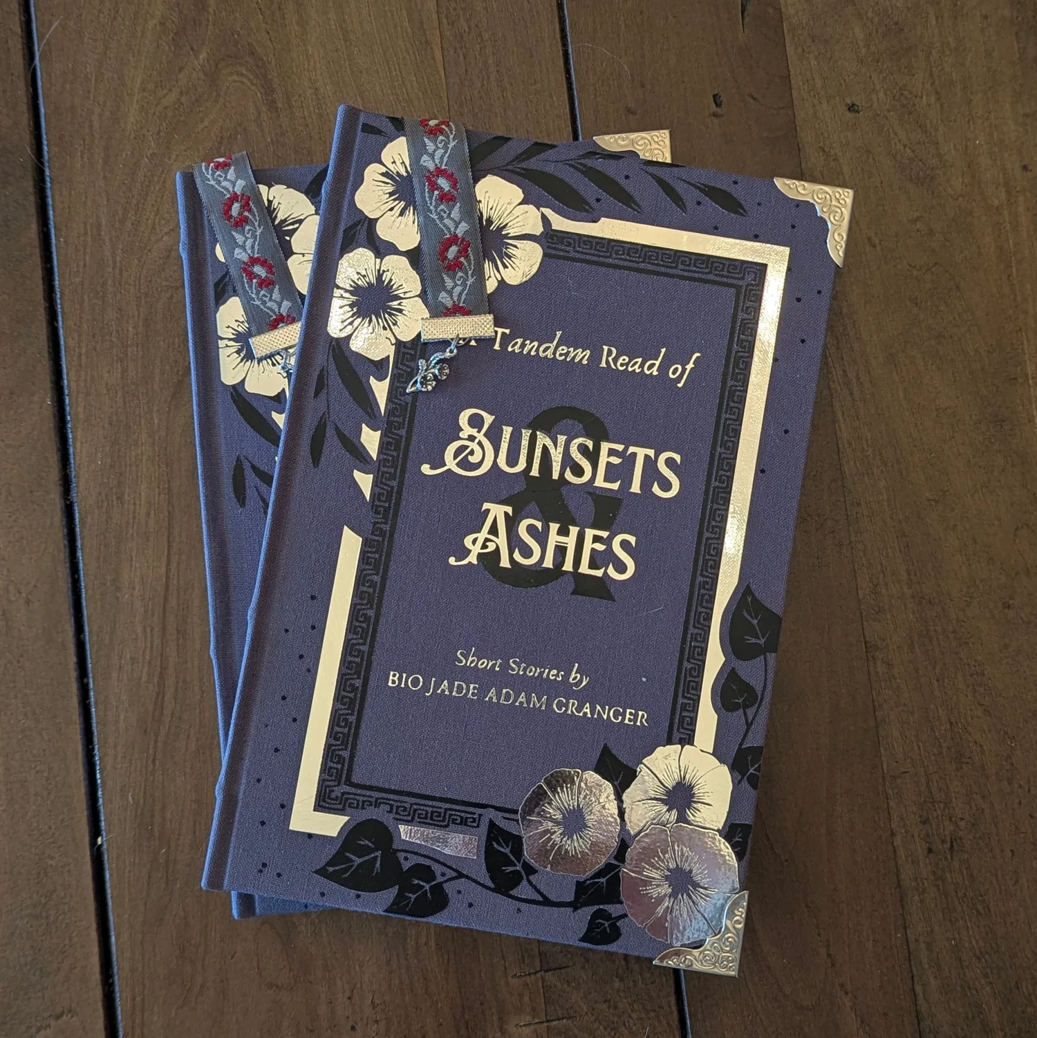 Two copies of Sunsets and Ashes stacked on dark wood: gray bookcloth with a silver and black HTV cover design, silver corner protectors and a jacquard ribbon marker.