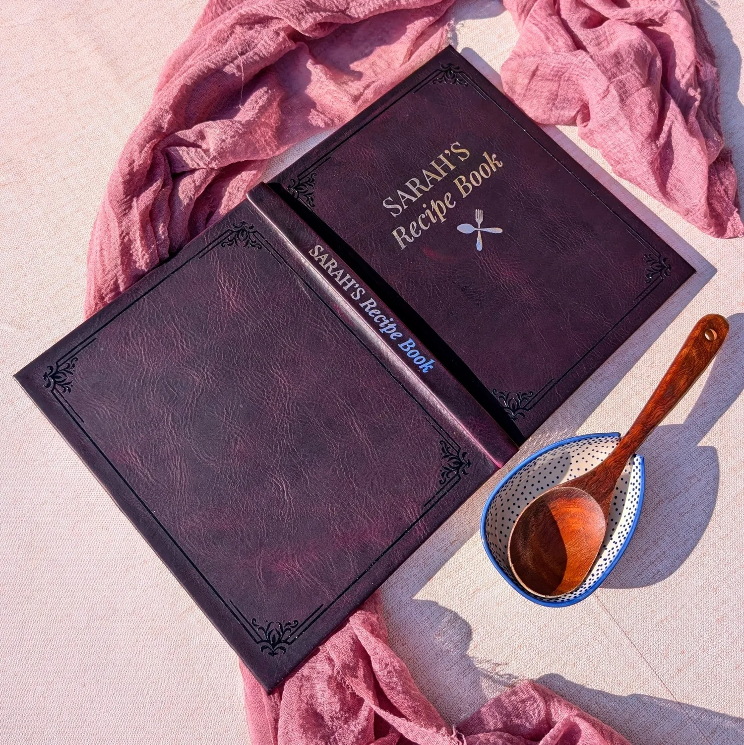 Sarah's Recipe Book in dark oxblood leather with a black HTV border and silver HTV title and spine, photographed beside a wooden spoon.