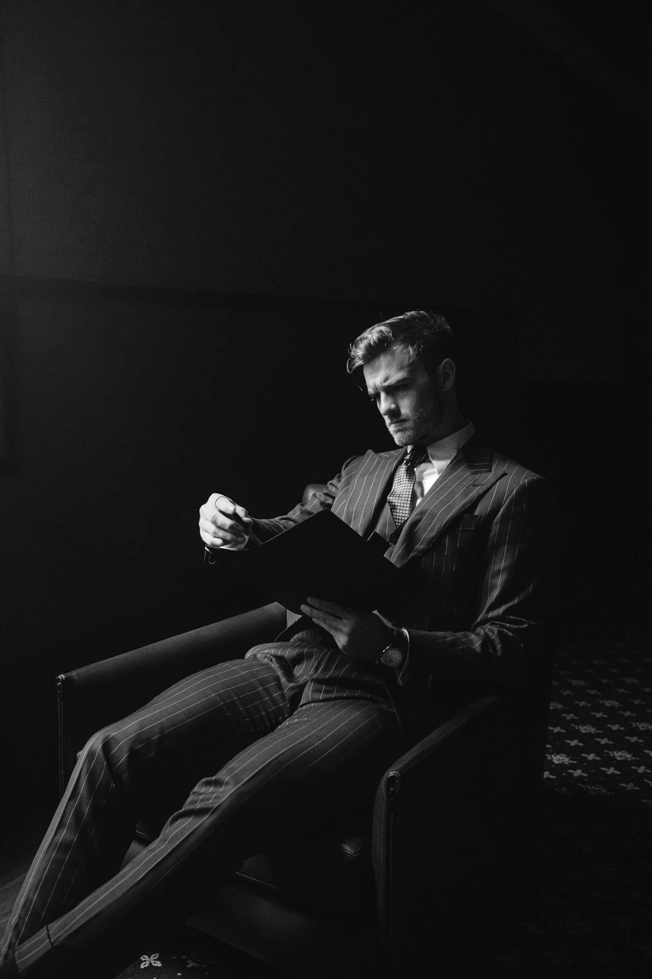 A man in a suit sitting back in a leather chair in low light
