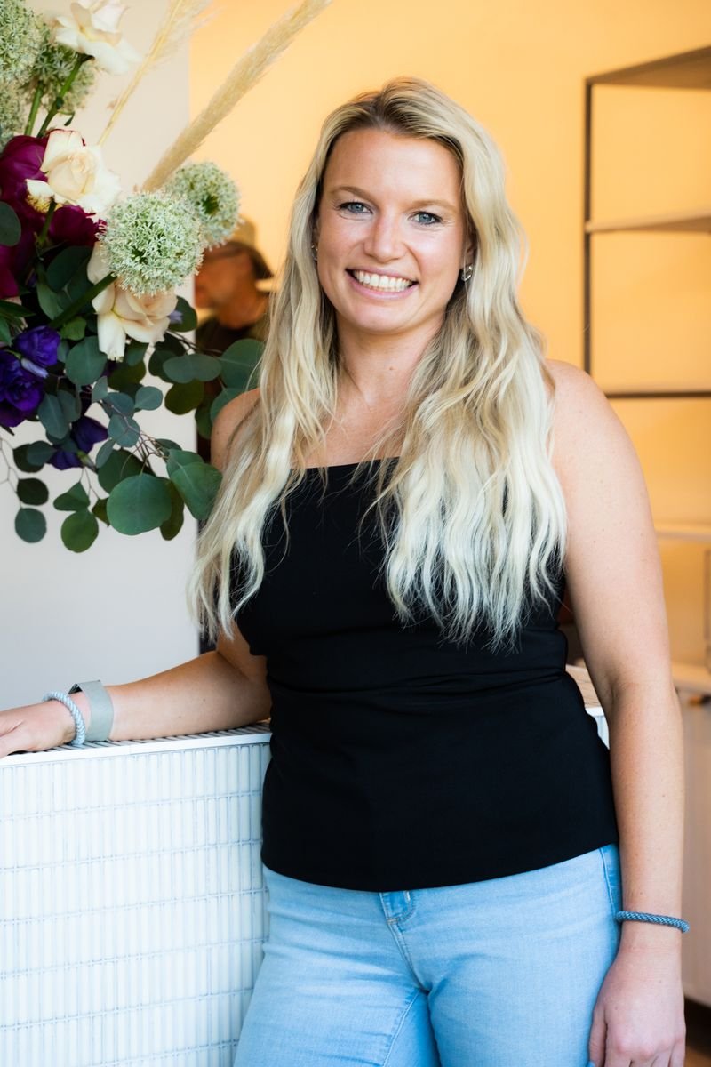 Headshot of Katherine Carder. She is leaning against the receptionist desk, looking at the camera, and smiling. Katherine has long blonde hair, tan skin and blue eyes. She is wearing a black tank top.