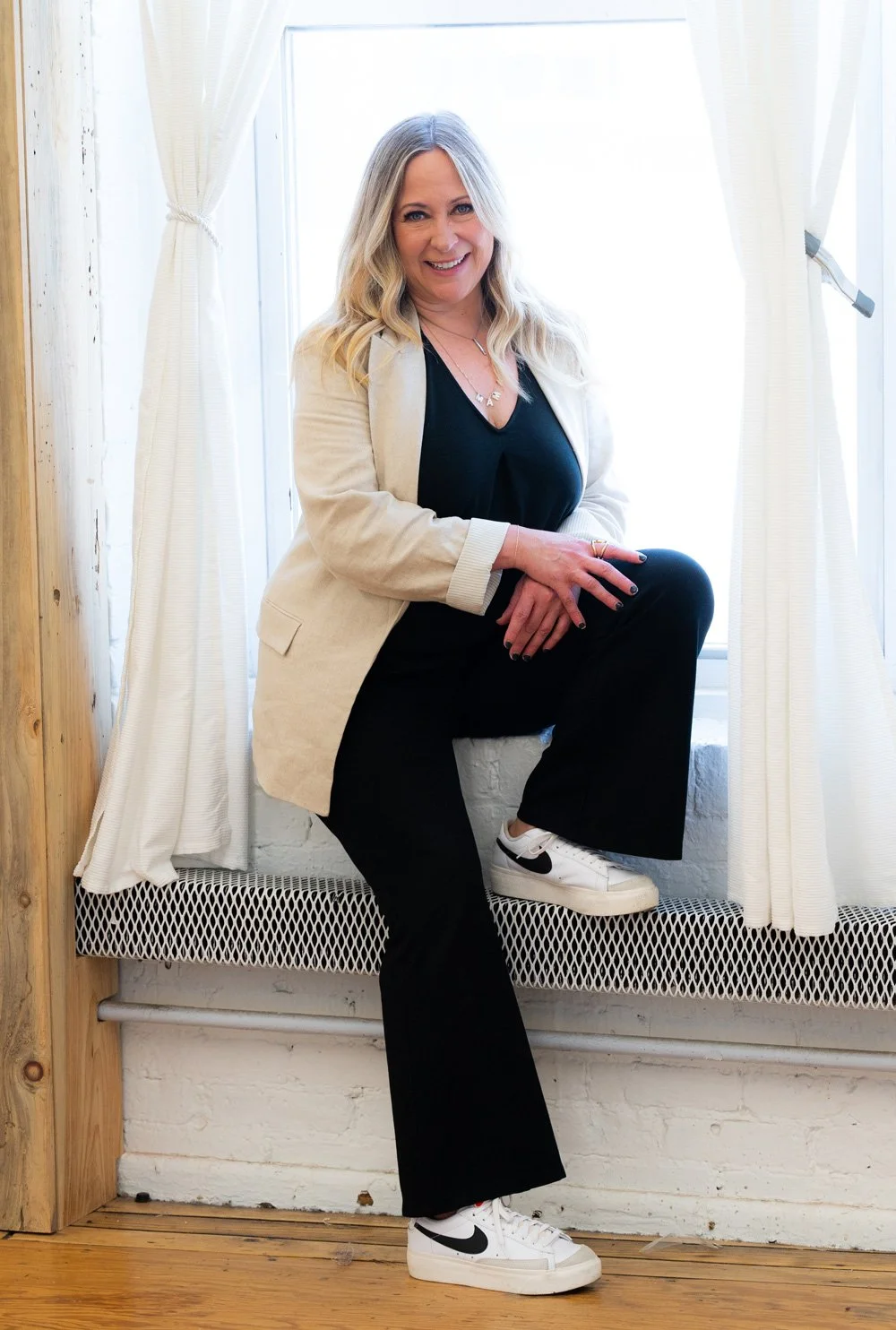 Headshot of Laurene Overlan. She is sitting on a windowsill with her hands on one knee. Laurene is wearing all black with a tan over coat. She has long, wany blond hair and is staring at the camera and smiling.