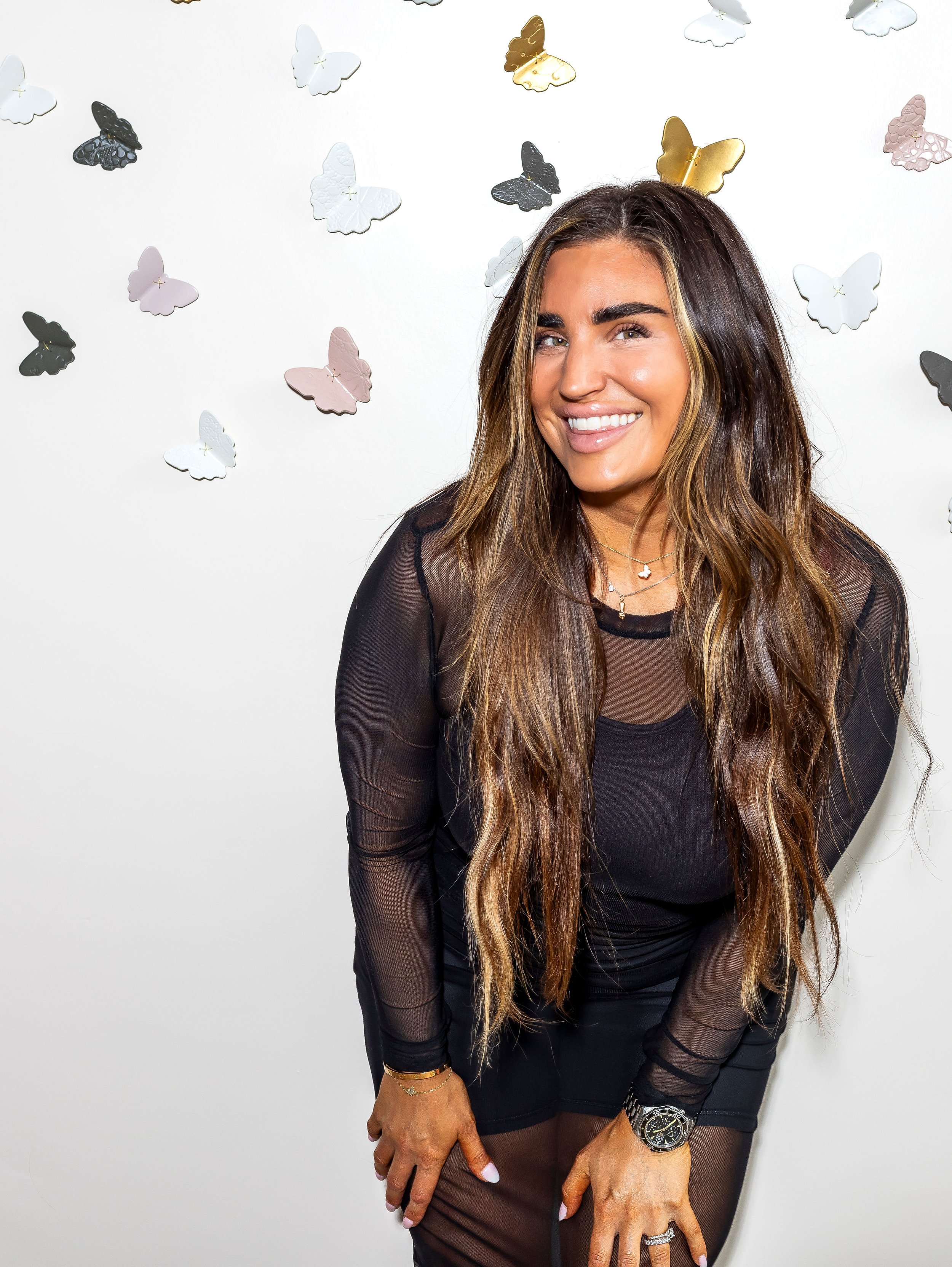 A woman standing in front of a grey wall with an array of butterflies adorning the wall. She has long, highlighted brown hair and is smiling while looking straight at the camera