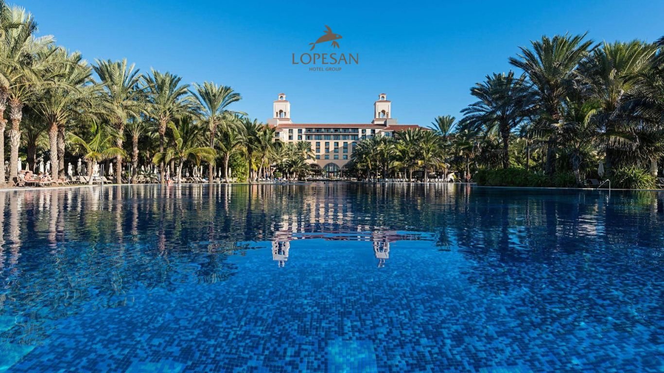 A serene resort view featuring the Lopesan Hotel Group’s pool area, surrounded by lush palm trees with the elegant hotel structure in the background under a clear sky.