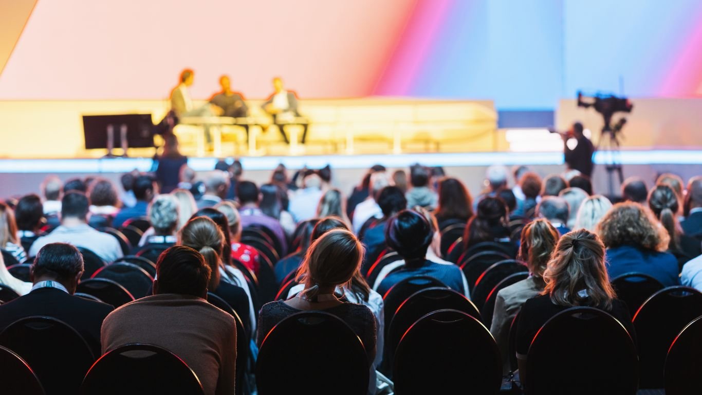 A conference scene with an engaged audience and a panel discussion on stage, highlighted by a camera operator and vibrant stage lighting.