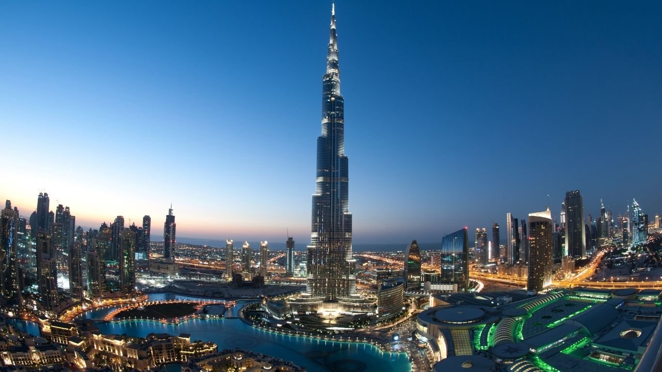 Panoramic view of Dubai’s skyline at dusk, featuring the Burj Khalifa and the city’s illuminated buildings against a clear sky.