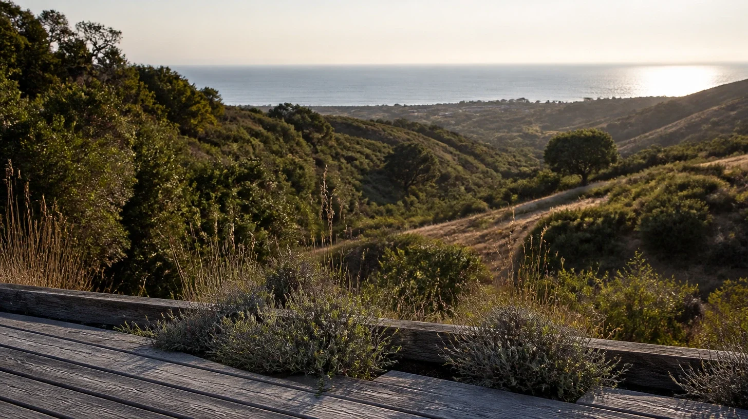 A weathered timber deck at a Malibu hillside property with coastal sage at its edge, the land falling away through chaparral and the ocean flat and silver along the far edge in late light