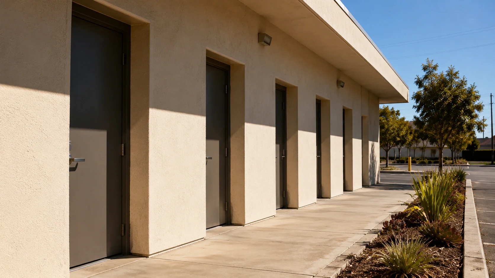 The shared exterior walkway of a small multi-tenant commercial building, with five identical unmarked tenant doors receding in a row under raking sunlight.