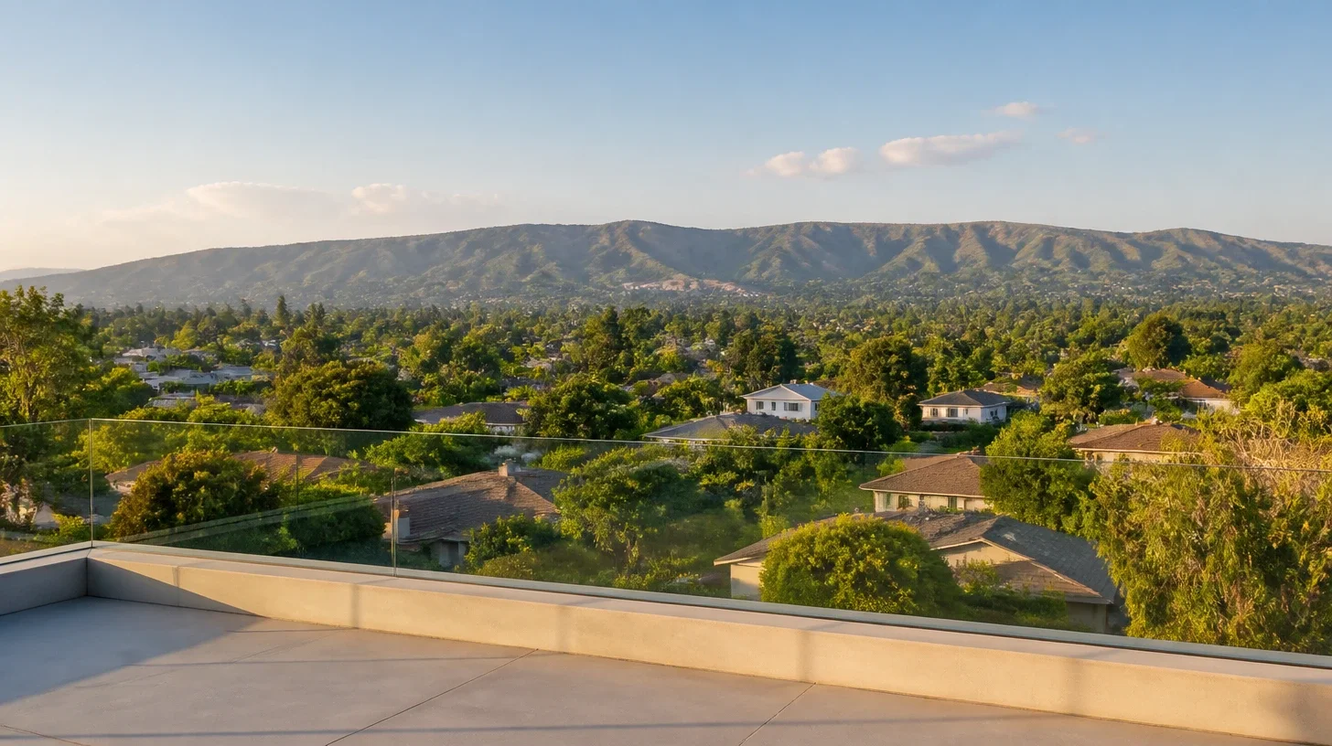 A wide view across Whittier from a terrace with a frameless glass balustrade, low rooftops and mature tree canopy filling the middle distance and the long ridge of the Puente Hills across the horizon in late afternoon light