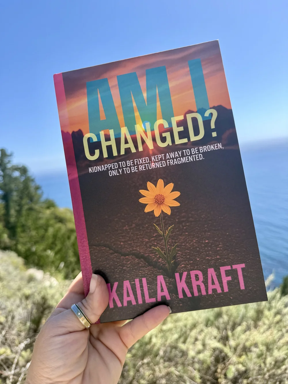 Kaila Kraft holding a copy of Am I Changed? above the Big Sur coastline