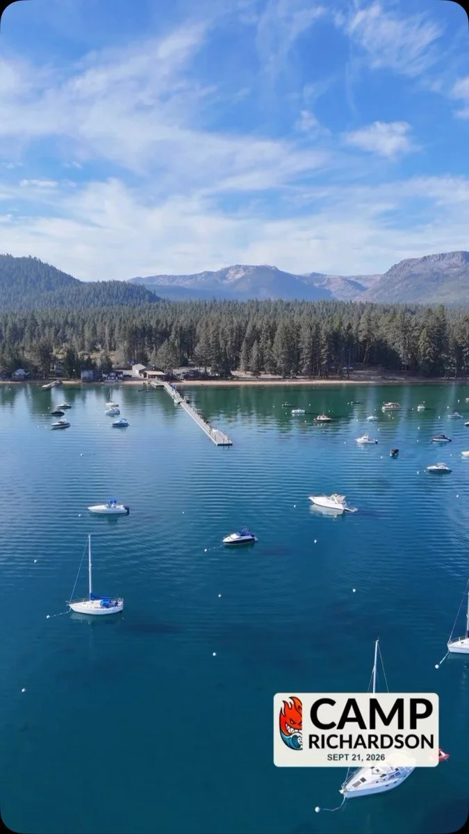 Boats moored in a Lake Tahoe cove beneath forest, mountains and high clouds.