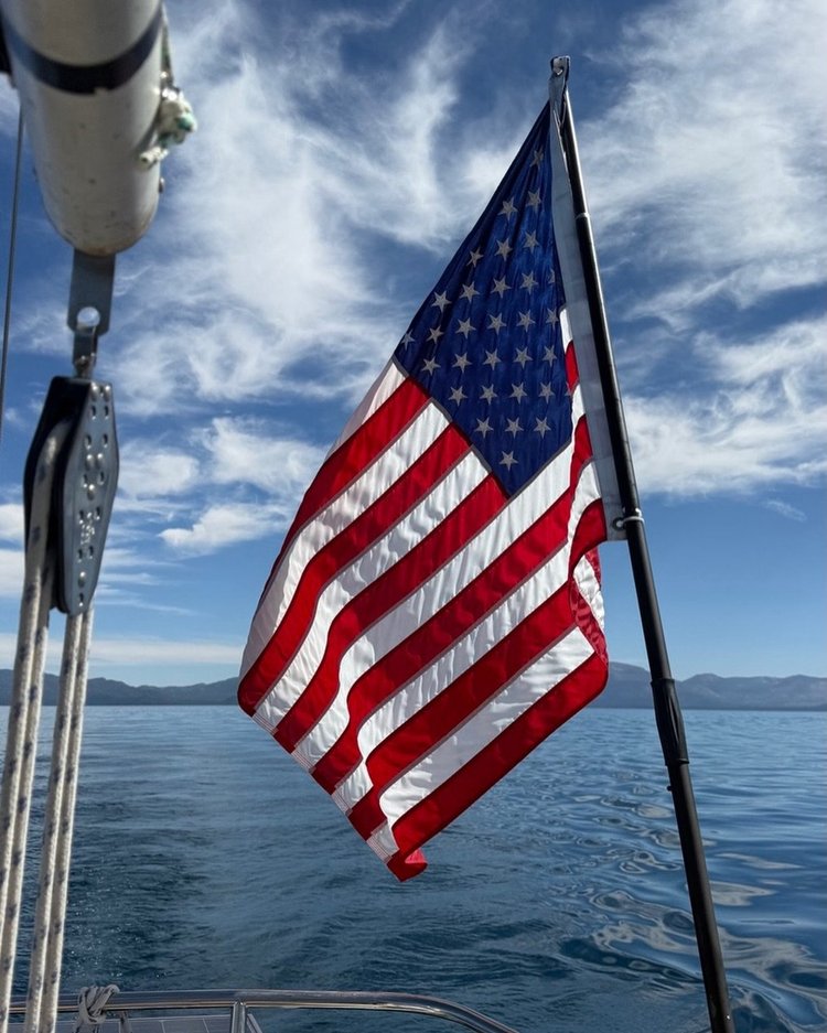 An American flag flies from a sailboat above Lake Tahoe, with mountains across the water.