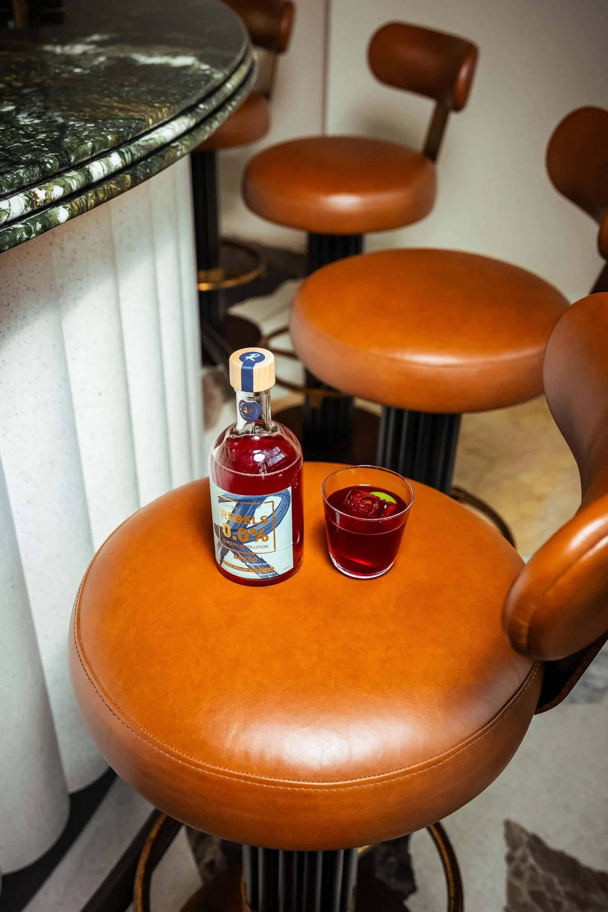 A bottle of Rebels 0.0% Rosso and a poured cocktail on a leather bar stool