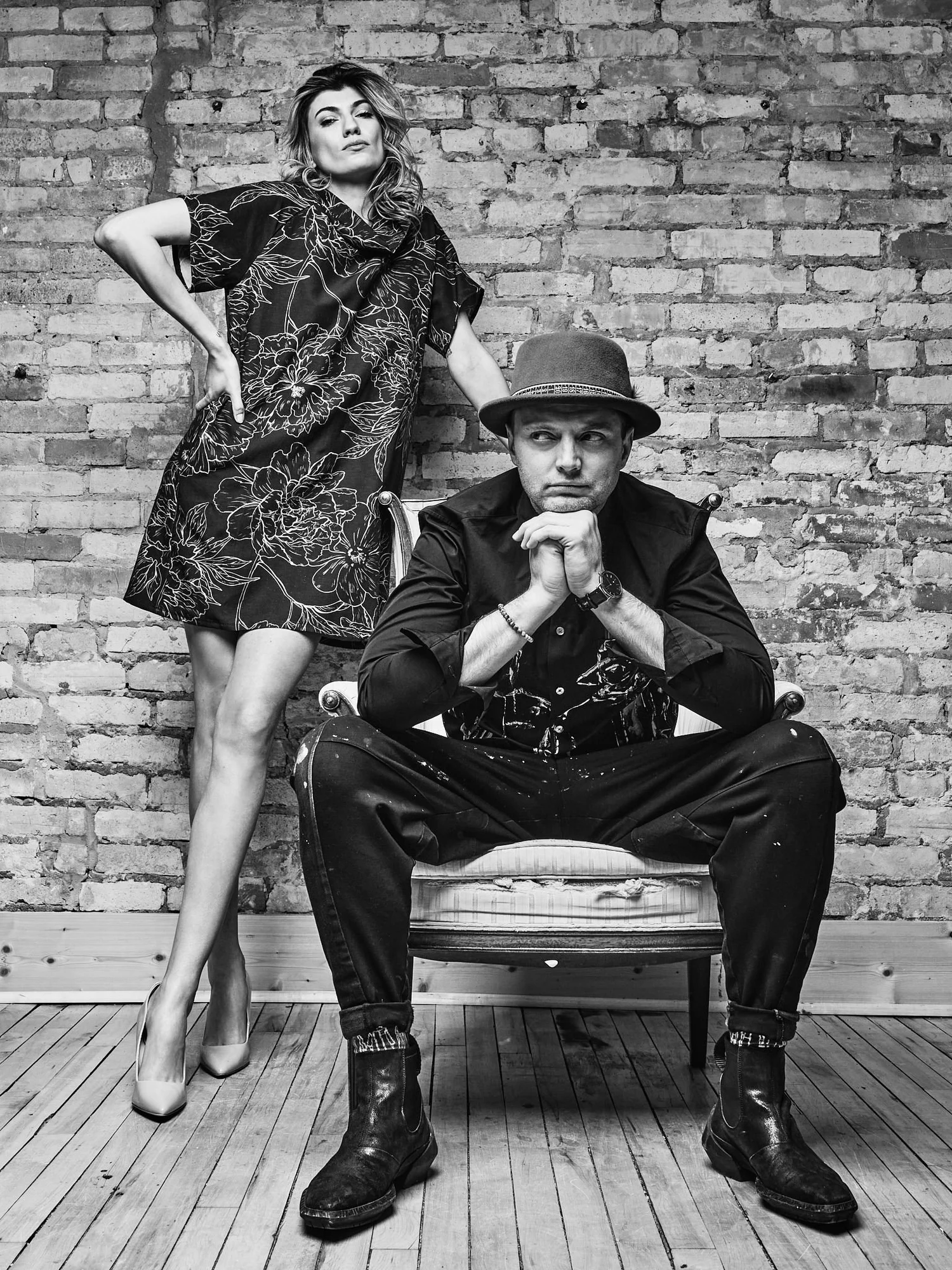 Designer Lennard Taylor seated in a hat beside a model in a patterned dress against a brick wall, black and white
