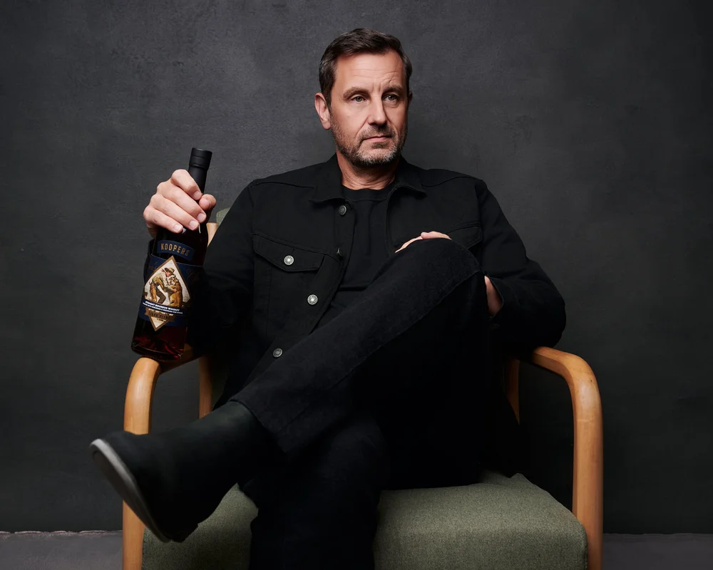 One of the owners of Koopers Whiskey seated in a wooden armchair, holding a bottle of the whiskey, for the brand's campaign