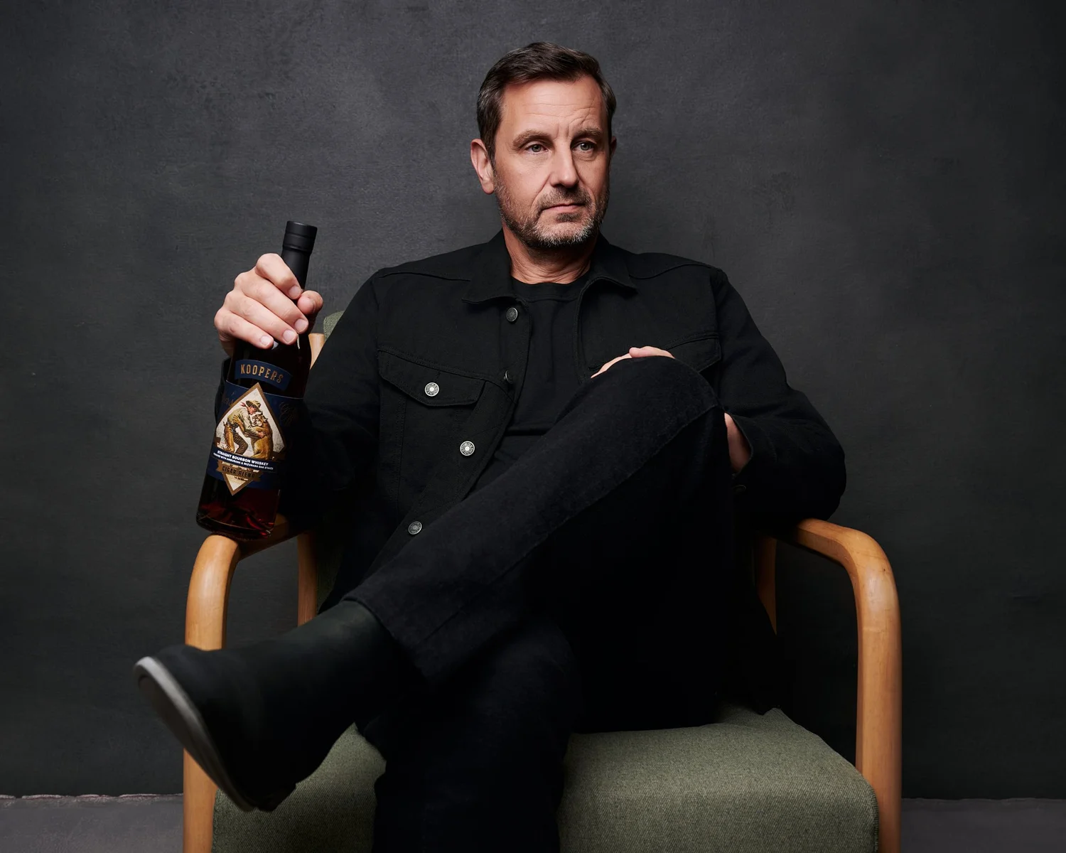 One of the owners of Koopers Whiskey seated in a wooden armchair, holding a bottle of the whiskey, for the brand's campaign