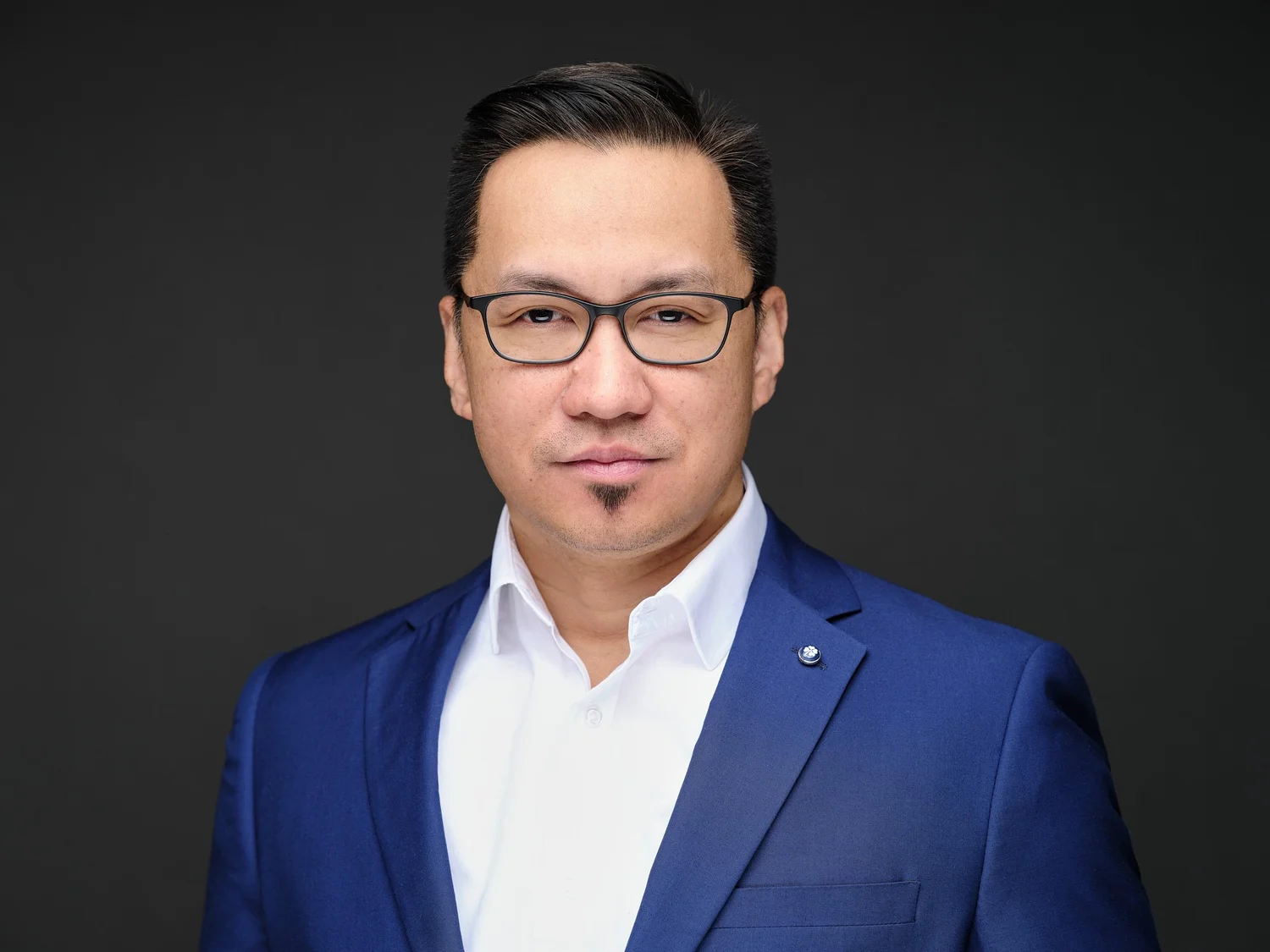 Color headshot of a man in a blue suit and white shirt on a dark ground, photographed by Ryan Dunbar