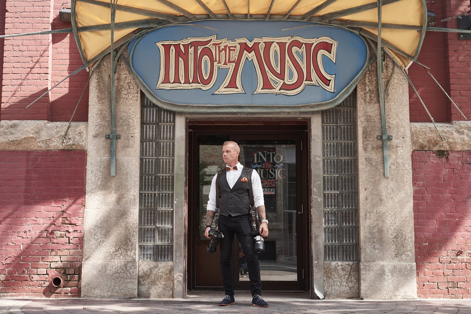 A photographer in a waistcoat standing in a shop doorway with two cameras on an Ikonic Leather Works harness
