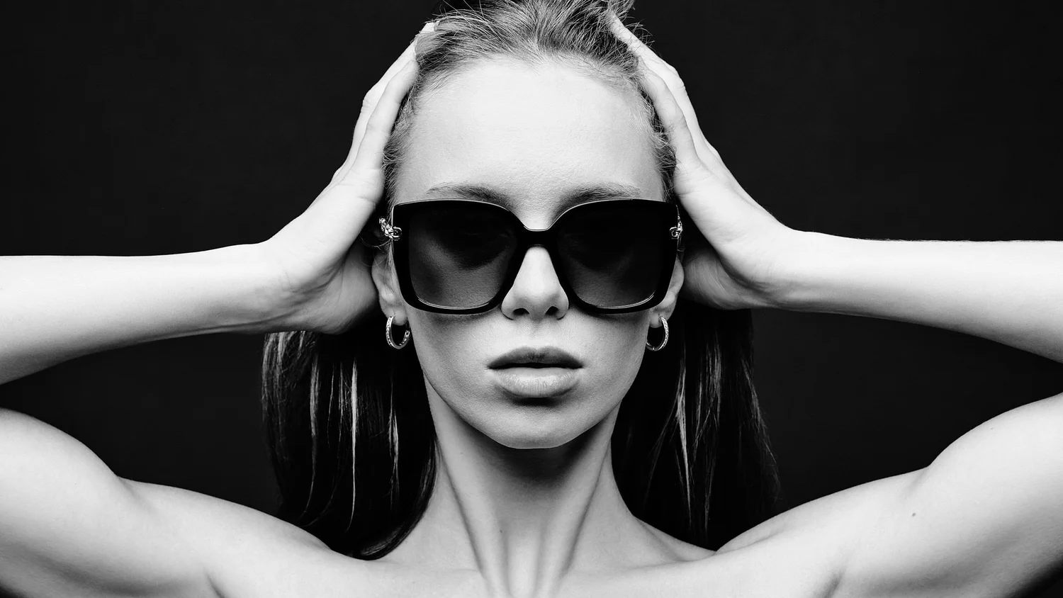 Model in dark sunglasses, hands on her head, black and white studio portrait for Village Optical