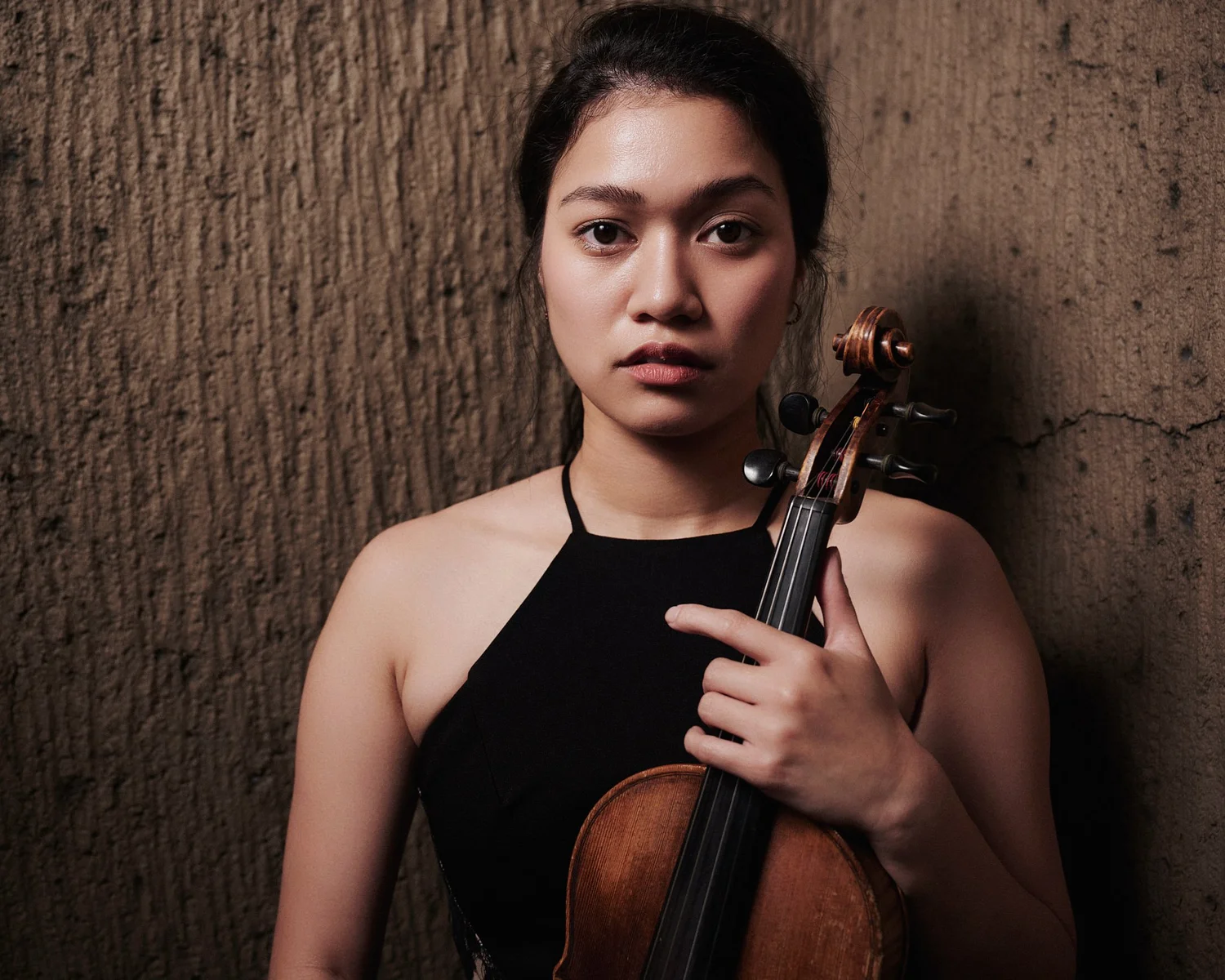 Eliza holding her violin against a textured wall, looking into the camera