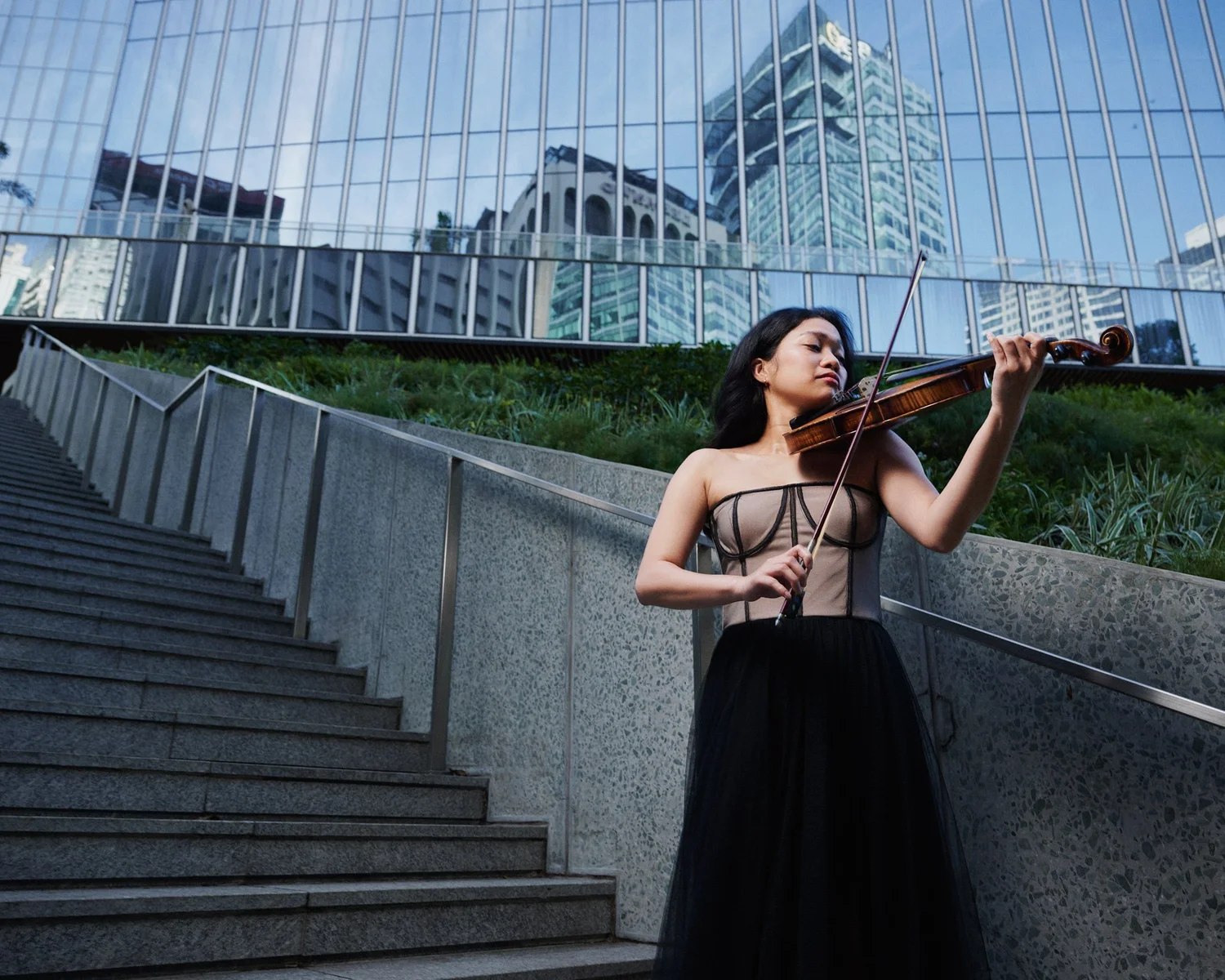 Eliza playing violin on concrete steps below a glass building