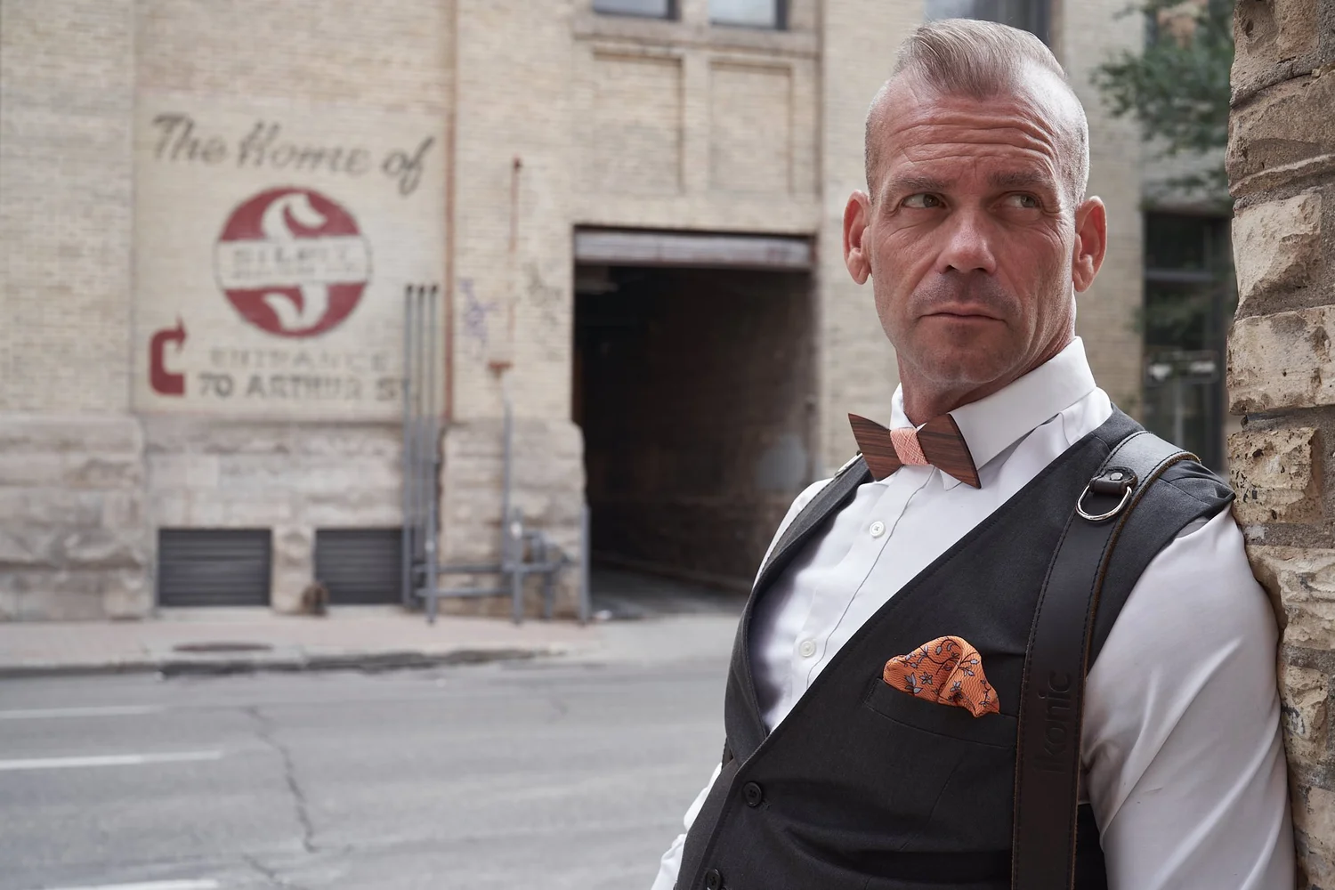 A photographer in a waistcoat and bow tie wearing an Ikonic Leather Works shoulder strap on a city street