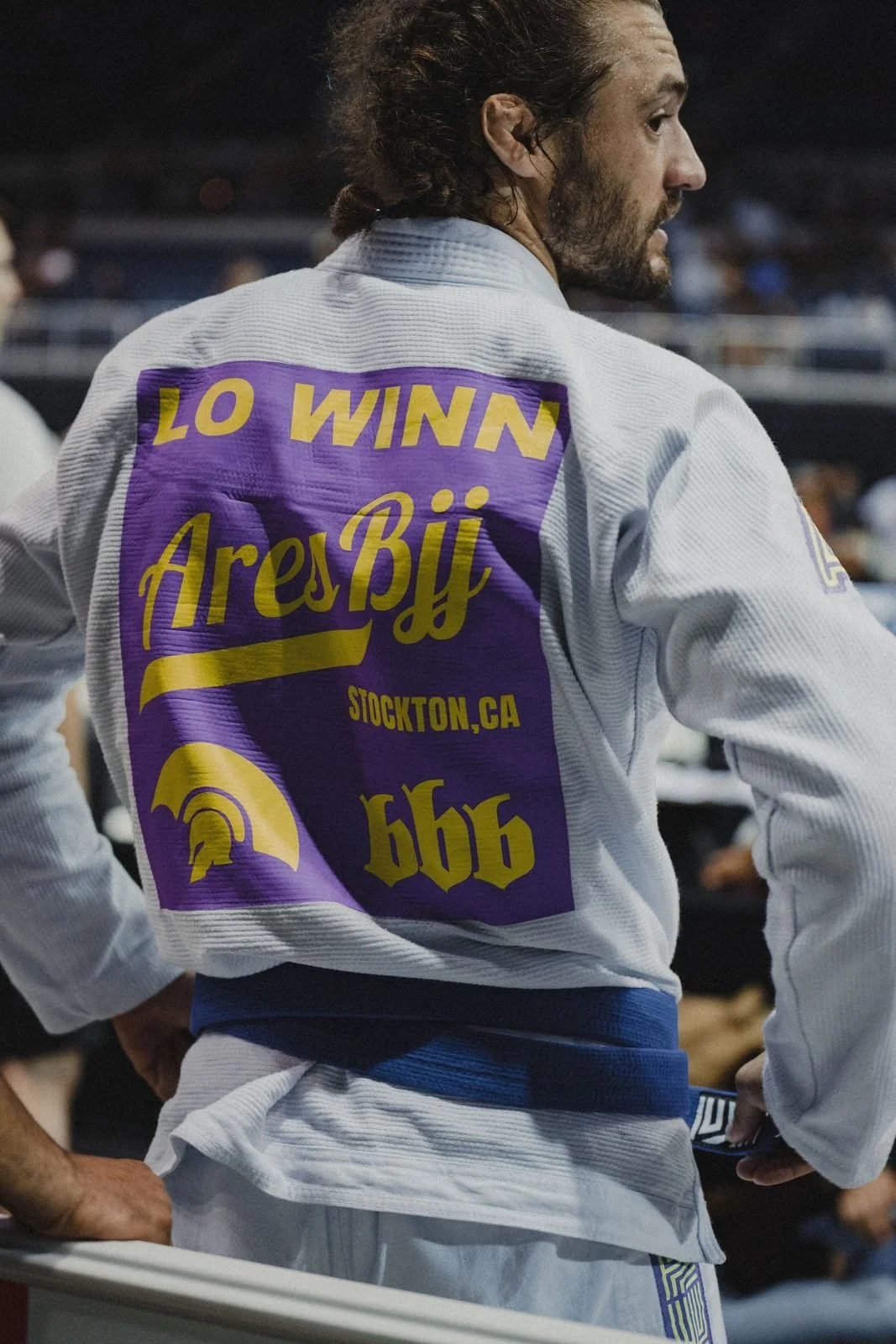 Logan from behind in a white gi with LO WINN on the back, walking out to compete